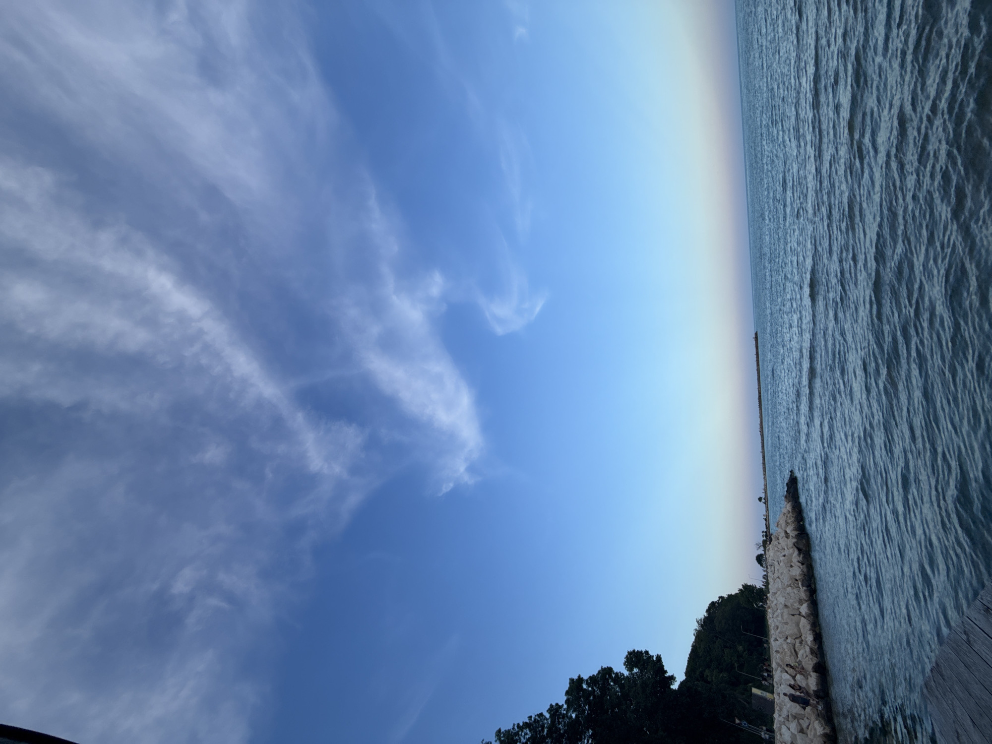 The image depicts a serene coastal scene likely taken at sunset or twilight in Varna, Bulgaria. The foreground features a wooden pier or boardwalk leading towards the water. The sea is calm with gentle ripples reflecting the soft hues of the sky. To the left, a rocky embankment lined with trees provides a natural border to the coastline. A few individuals are visible on this embankment, appearing to be enjoying the view. Further in the distance, a breakwater extends into the sea, with a faint silhouette of structures at its end. The sky above is a gradient of deep blue at the top, transitioning to lighter shades with wispy clouds towards the horizon, suggesting the fading light of day. The overall atmosphere is tranquil and picturesque.