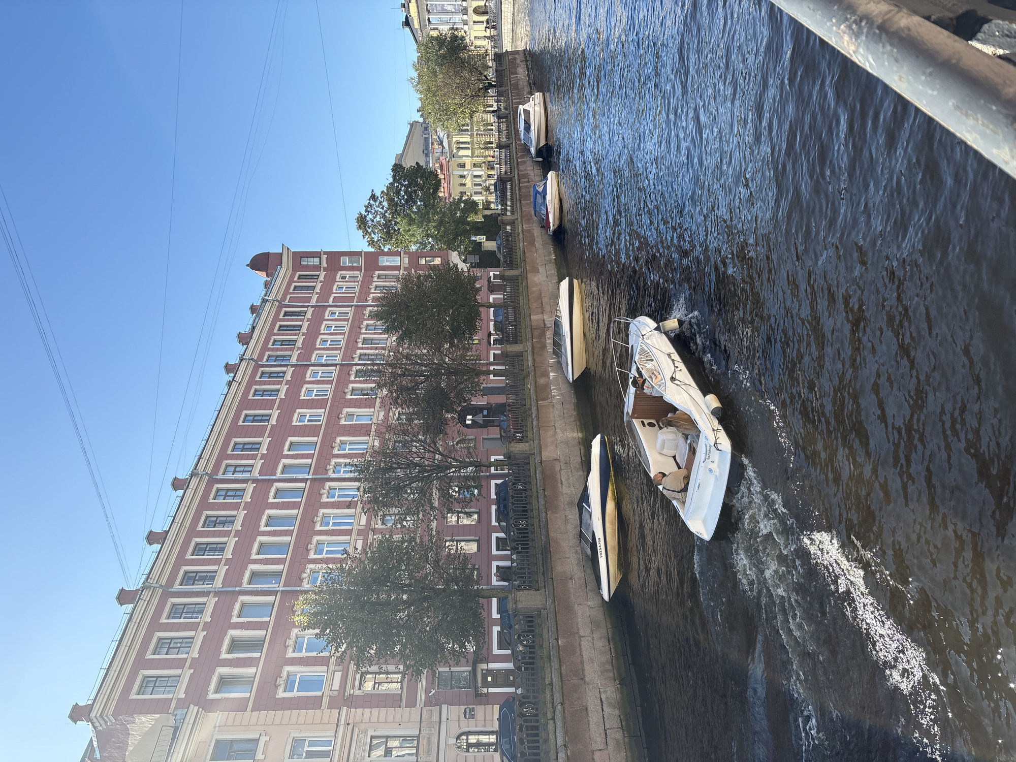 The image depicts a sunny day in Saint Petersburg, Russia, specifically along a canal. A multi-story building with a reddish-pink facade and numerous windows lines the left bank of the canal. Several trees with green foliage are situated between the building and the canal. The canal itself is populated with multiple boats. In the foreground, a white motorboat with a canopy is moving away from the viewer, leaving a wake in the water. Further down the canal, there are at least four other motorboats moored or moving. The water in the canal reflects the blue sky and surrounding buildings. The right side of the image shows a railing. The sky is clear and blue.