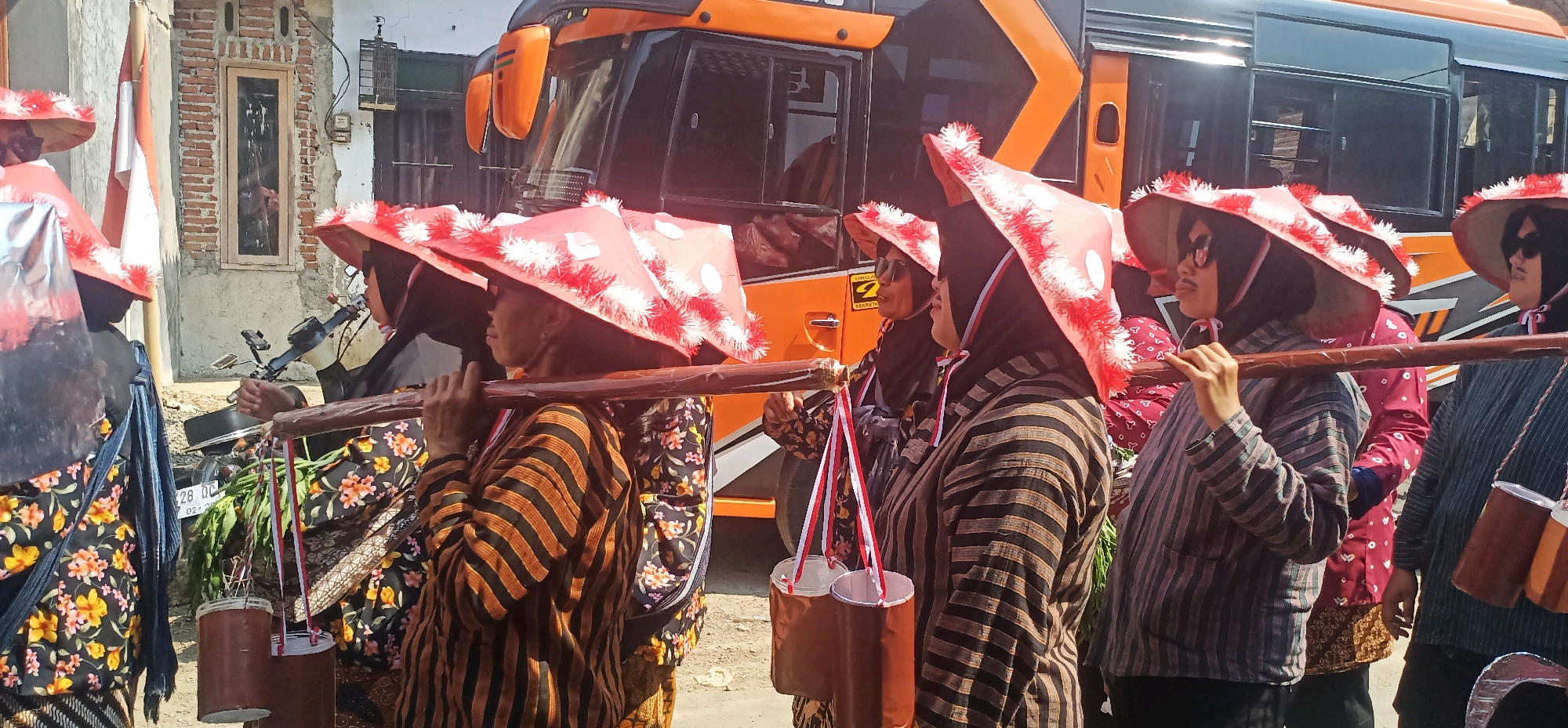 In the city of Kincang, Indonesia, a group of people, primarily women, are participating in what appears to be a cultural procession or event. They are dressed in traditional attire, featuring striped long-sleeved shirts and patterned skirts or sarongs. Many of the women are wearing conical hats adorned with red and white fringes and white polka dots. They are carrying wooden poles across their shoulders, from which are suspended buckets or containers, some with green foliage. The background reveals a brick building with a window and a large orange and black bus, suggesting an urban or semi-urban setting. The overall atmosphere seems festive and communal, with individuals engaged in carrying items as part of a shared activity.