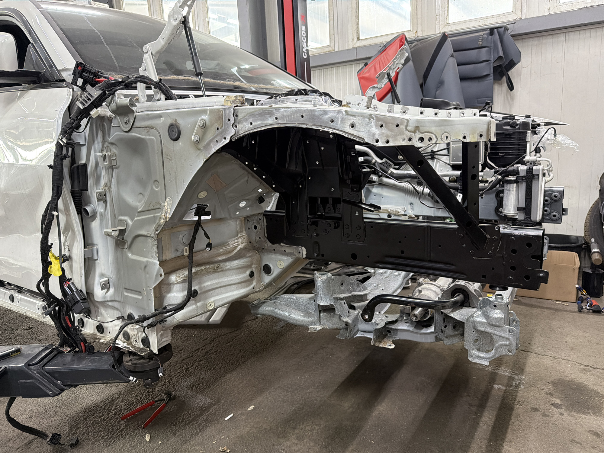 The image shows the front section of a car chassis that has been partially disassembled, likely in a repair shop. The car's white exterior is visible on the left, with the underlying metal structure exposed. Various wires and electrical components are attached to the chassis, some bundled together with black tape. A black metal frame is prominently featured in the center, supporting parts of the vehicle. Various tools, including a pair of red-handled pliers, are scattered on the concrete floor, suggesting ongoing work. In the background, there are windows and a large red and black structure that appears to be a car lift. The overall environment appears to be an indoor workshop. The scene is forensic in its detail, presenting the disarray of a vehicle undergoing repair without any embellishment or interpretation of the state of the car. The image is a factual representation of mechanical disassembly.