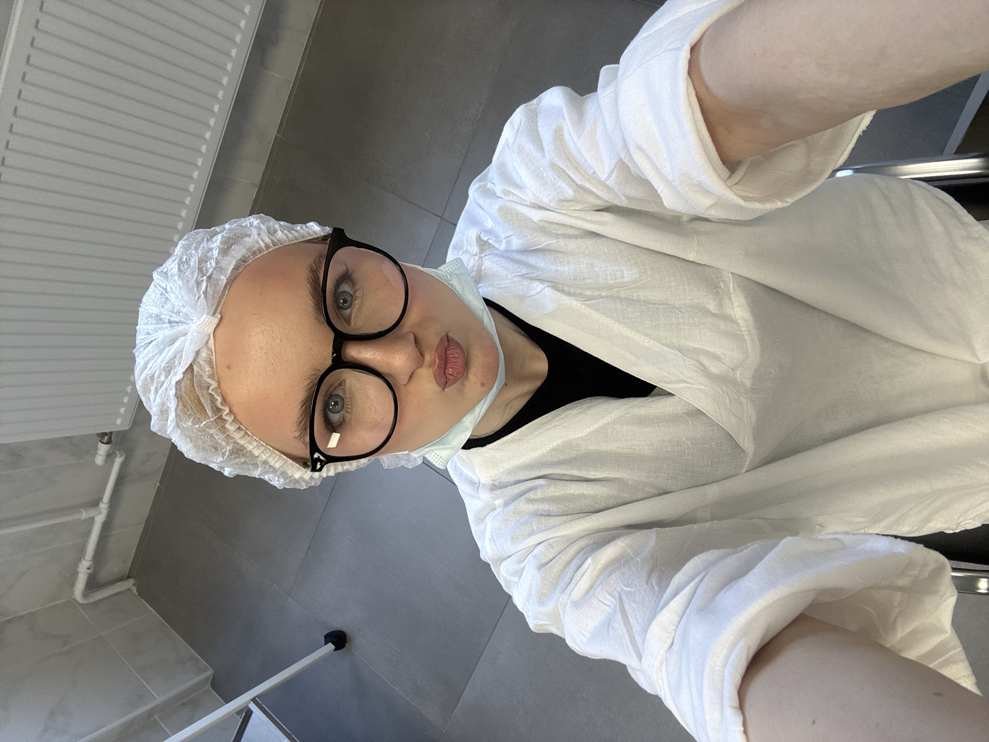 A woman wearing a white hairnet, black-rimmed glasses, a light blue surgical mask hanging below her chin, and a white robe is taking a selfie in an indoor setting. She has her lips pursed and is looking directly at the camera. The background shows grey tiled flooring, white pipes, and a white radiator. There is a hint of a white chair or stool.