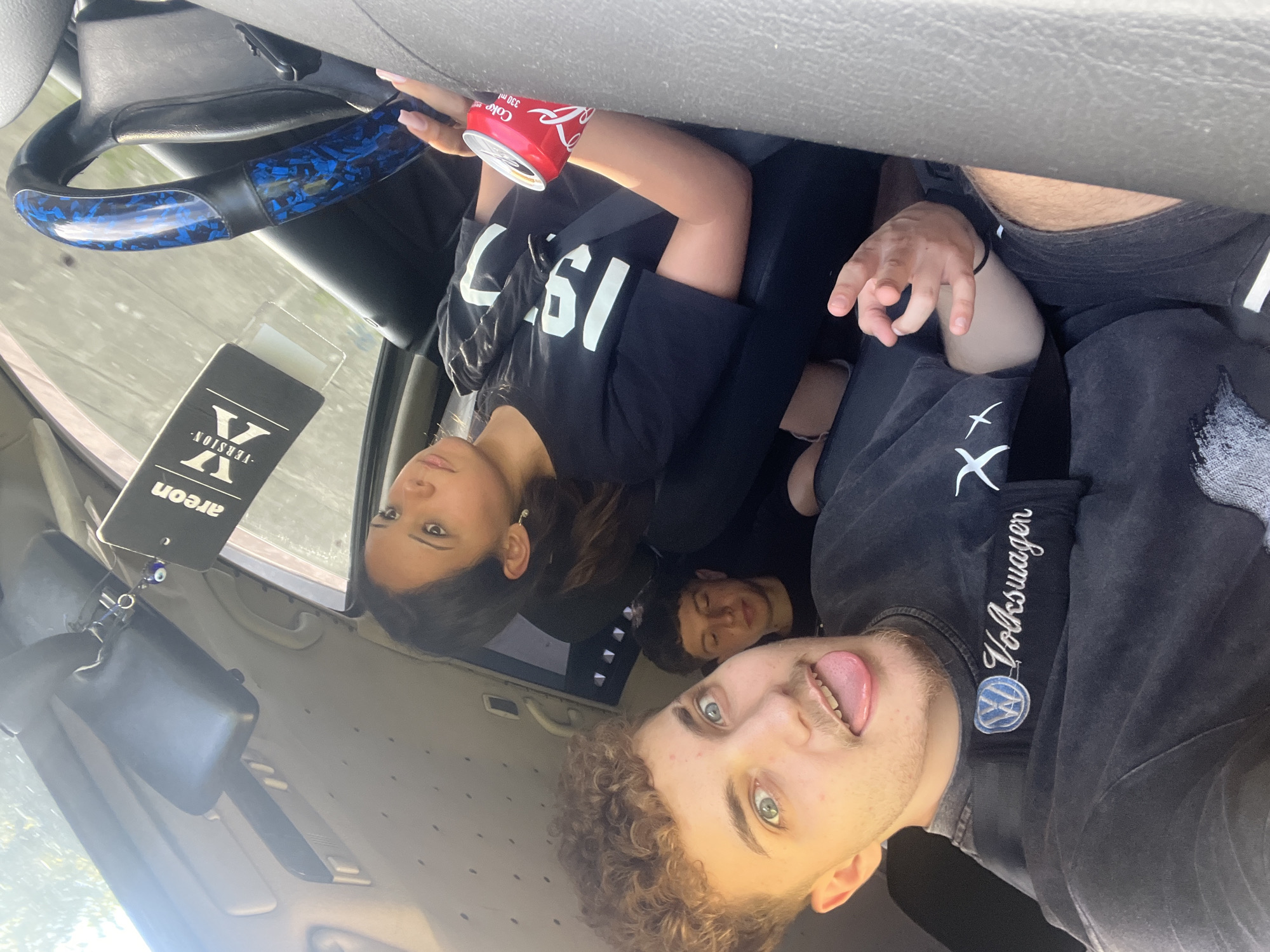 The image shows four young people in a car. The person in the foreground, on the left, is taking the selfie. They have curly brown hair and are sticking their tongue out. They are wearing a dark t-shirt with a Volkswagen logo on it. To their right, a person with their eyes closed is leaning on the seat. Further to the right, a young woman with dark hair is visible, wearing a black t-shirt with the number "1997" on it. Her left hand is resting on a red can of Coca-Cola. Another person's arm is visible on the far right. The car's interior is visible, including a steering wheel with a blue and black patterned cover and an air freshener hanging from the rearview mirror. The scene appears to be in Vlahovo, Bulgaria, as indicated by context provided with the image. The lighting suggests it is daytime.