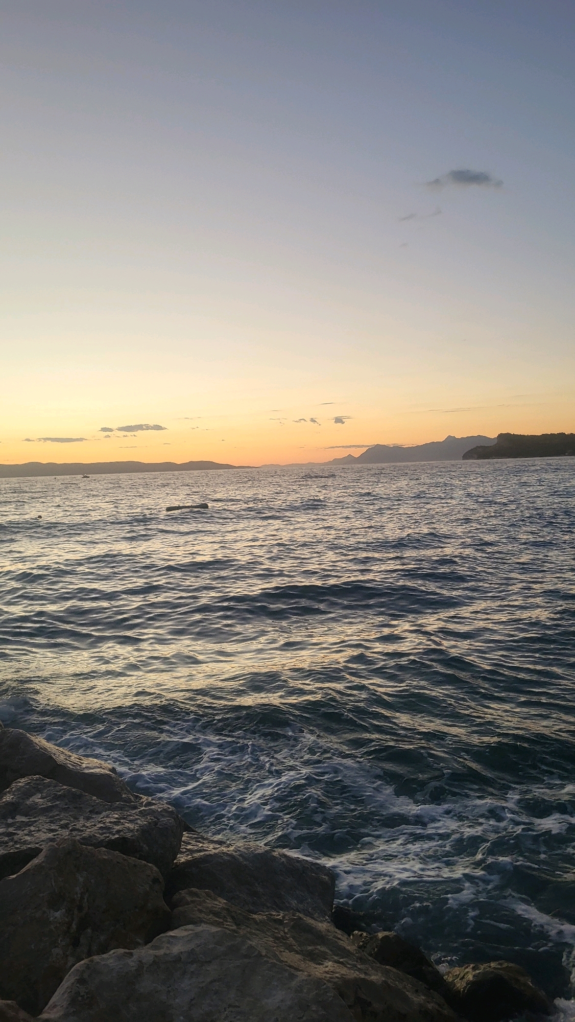The image depicts a serene coastal scene at dusk, likely in Općina Tučepi, Croatia. The foreground is dominated by large, grey rocks that form a natural breakwater. Beyond the rocks, the deep blue water of the sea is captured with gentle waves creating white foam along the shore. In the middle ground, a dark, cylindrical object, possibly a log or debris, floats on the surface of the water. Further in the distance, the calm sea stretches towards a low-lying coastline characterized by hills and mountains under a gradient sky. The sky transitions from a pale blue at the top to soft hues of orange and yellow near the horizon, indicating the sun has recently set or is about to rise. A few small clouds are visible in the upper portion of the sky. The overall atmosphere is peaceful and tranquil.