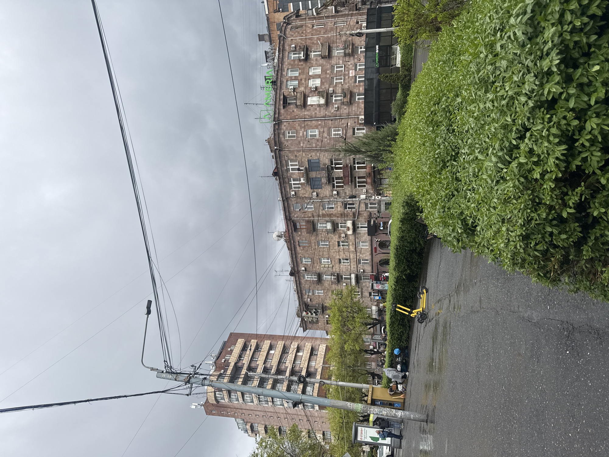 An urban street scene unfolds in Yerevan, Armenia, under a heavily overcast, grey sky, suggesting gloomy weather or recent rain, evidenced by the wet asphalt road. The image captures a cross-section of city life, featuring prominent buildings, street infrastructure, and scattered objects.
Dominating the left side of the frame are numerous overhead power lines and communication cables crisscrossing the sky, supported by a tall street light pole. Attached to this pole is a modern street lamp fixture, and near its base, a yellow trash receptacle or kiosk is visible, along with a small sign bearing Armenian text.
In the mid-ground, a large, multi-story building with a distinctive reddish-brown stone facade stands out. This building is characterized by many windows, some with small balconies, and numerous external air conditioning units. A large green sign spelling "AMERIABANK" is prominently displayed on its roof, and the name is also visible on a darker facade section lower down. Further in the background, another tall, lighter-colored residential or commercial building with multiple stories and windows can be seen.
Along the right side of the street, a dense, well-maintained green hedge runs, bordering what appears to be a sidewalk. Several yellow electric rental scooters are parked neatly near this hedge. A few indistinct human figures are visible on the sidewalk in the distance, with one person appearing to wear a light-colored poncho or raincoat, reinforcing the impression of wet conditions. The overall atmosphere is that of a typical, albeit damp, city day.