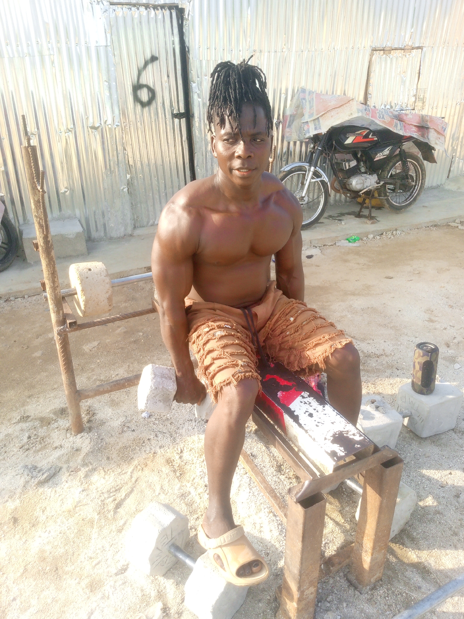 A muscular, bare-chested man with dreadlocks, wearing distressed tan shorts and tan slides, sits on a rusty weight bench outdoors. He is holding a concrete dumbbell in his right hand. The ground is covered in sand and gravel. Behind him, a corrugated metal structure stands, with a black spray-painted number "6" on it. To the right of the man, a black motorcycle with a white and red logo is parked, covered with what appears to be tarpaulin or bedding. The setting suggests a utilitarian outdoor gym in a possibly developing area, consistent with parts of Indo, Nigeria. The overall lighting indicates daylight.