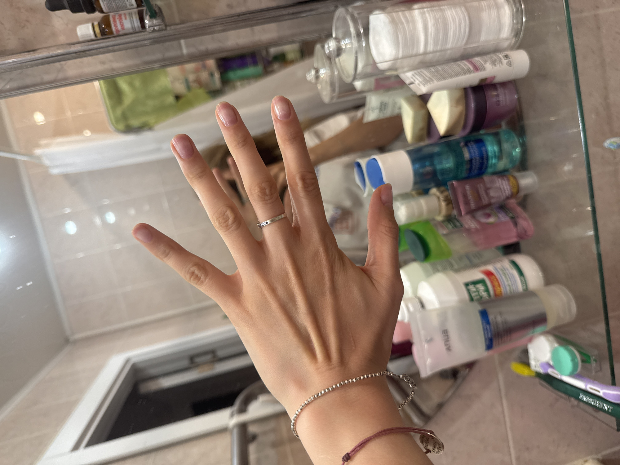 A hand is placed in front of a bathroom mirror. The hand has a silver ring on the ring finger and two bracelets on the wrist. The bathroom mirror has various items on its shelf, including cotton pads, creams, lotions, and toothbrushes. The background suggests a bathroom setting with light-colored tiles. There is no information in the image to indicate it is in Izhevsk, Russia.