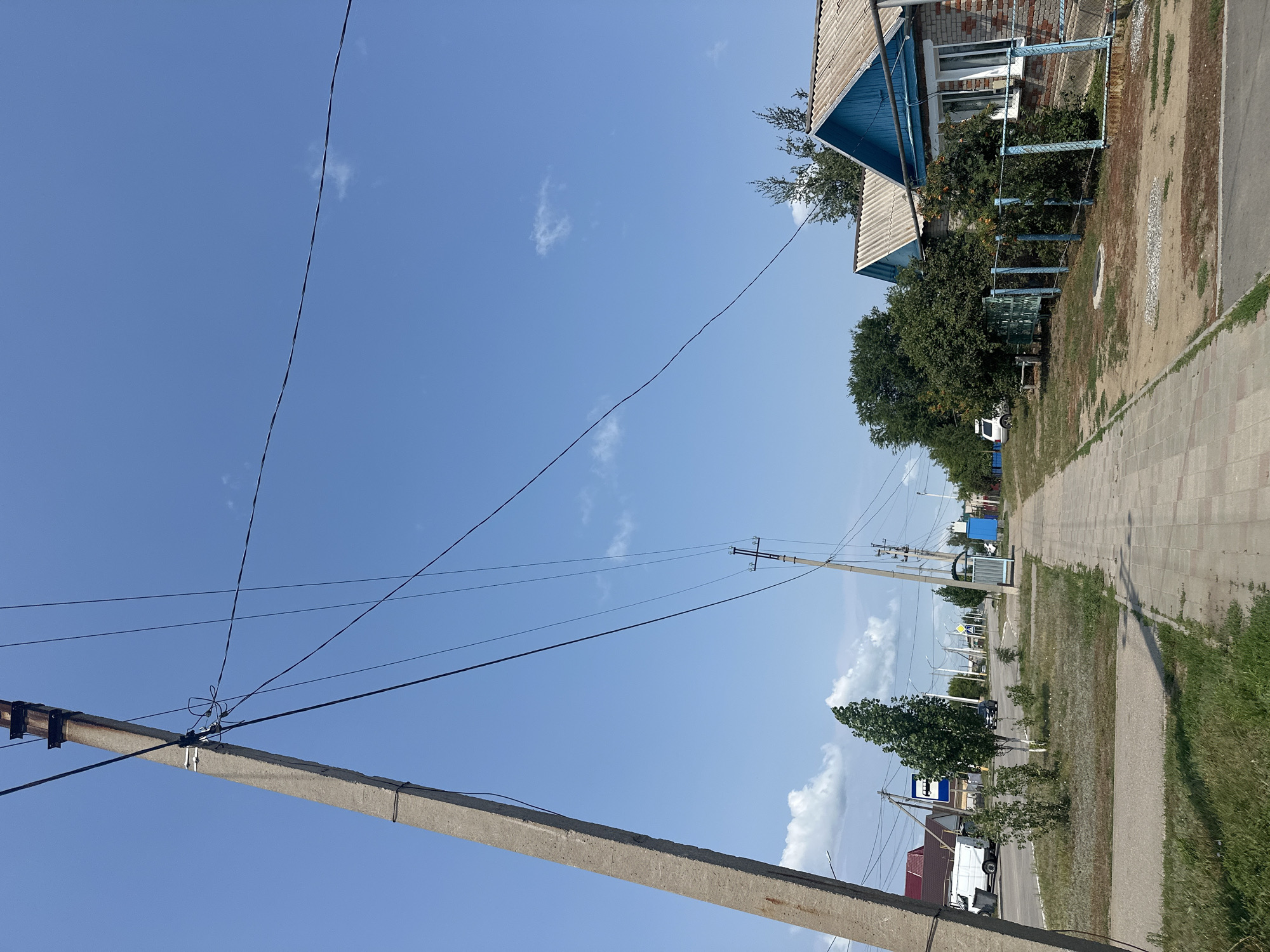 The image depicts a street scene in the city of Tobyl, Kazakhstan, on a clear, sunny day. In the foreground, a large concrete utility pole stands on the left side of the frame, leaning slightly. Several overhead wires extend from this pole, crisscrossing the blue sky. A paved sidewalk runs along the right side of the street, bordered by a grassy area.
To the right of the sidewalk, a brick house with a blue-painted wooden facade and a corrugated metal roof is visible. A light blue metal fence encloses a yard with some greenery and trees. Further down the street, more houses and trees can be seen.
On the left side of the street, across from the houses, there is a grassy median with several trees and bushes. A road is visible with a white van and a black car driving on it. A blue bus stop sign with a bus symbol is also present. In the distance, more utility poles and wires stretch along the street, suggesting a residential or semi-urban area. The overall environment appears to be an ordinary street in a town.