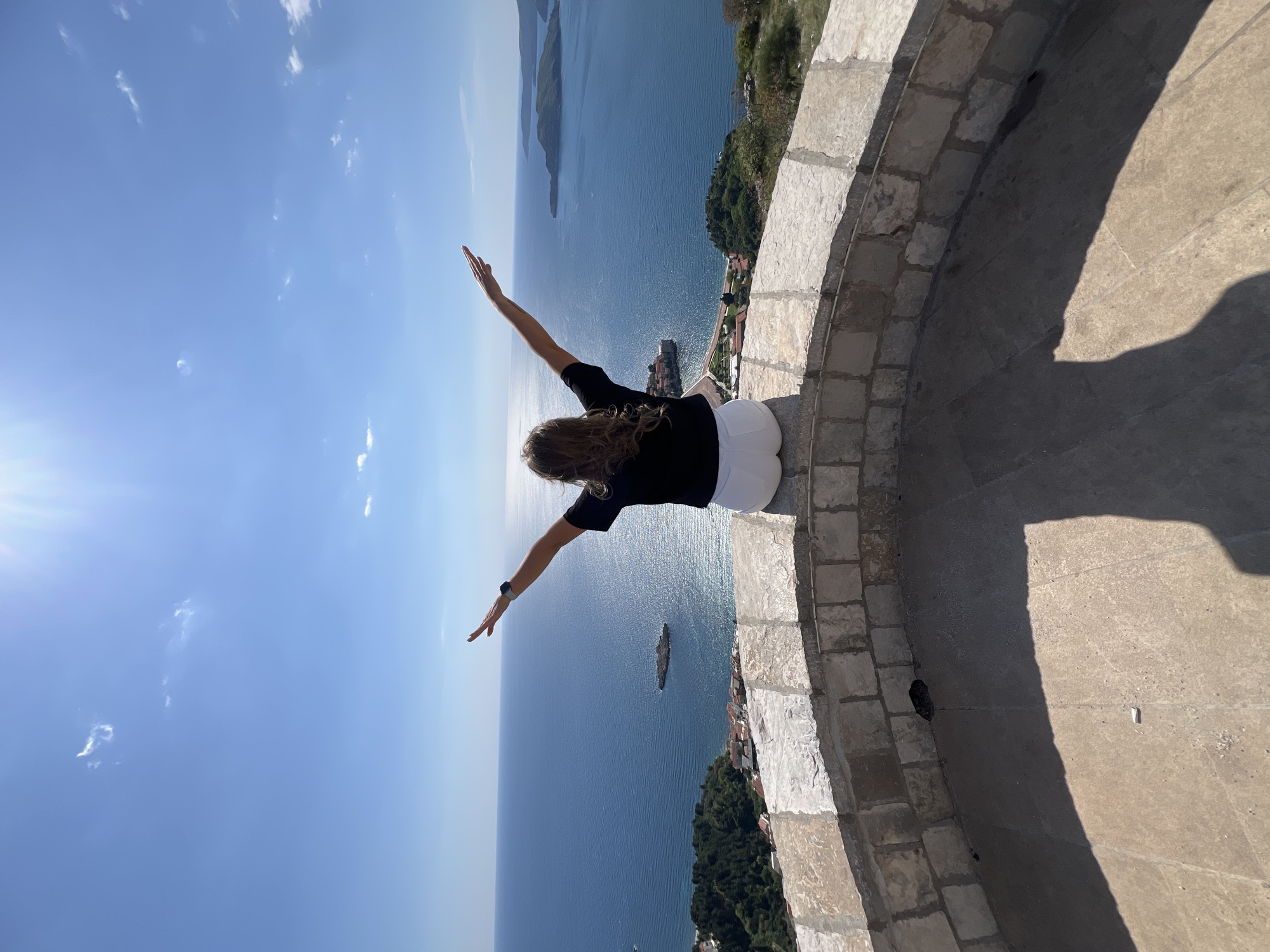 A woman sits on a stone wall with her back to the camera, arms outstretched, against a backdrop of blue sky and ocean. The location appears to be a high vantage point, possibly part of the medieval fortress of Sveti Stefan, overlooking the Adriatic Sea. The woman is wearing a black shirt and white shorts. She's sitting on the edge of a rounded stone barrier that has a small opening at the base. The shadow cast by the wall stretches across the paved ground. The sun is shining brightly, reflecting off the water.