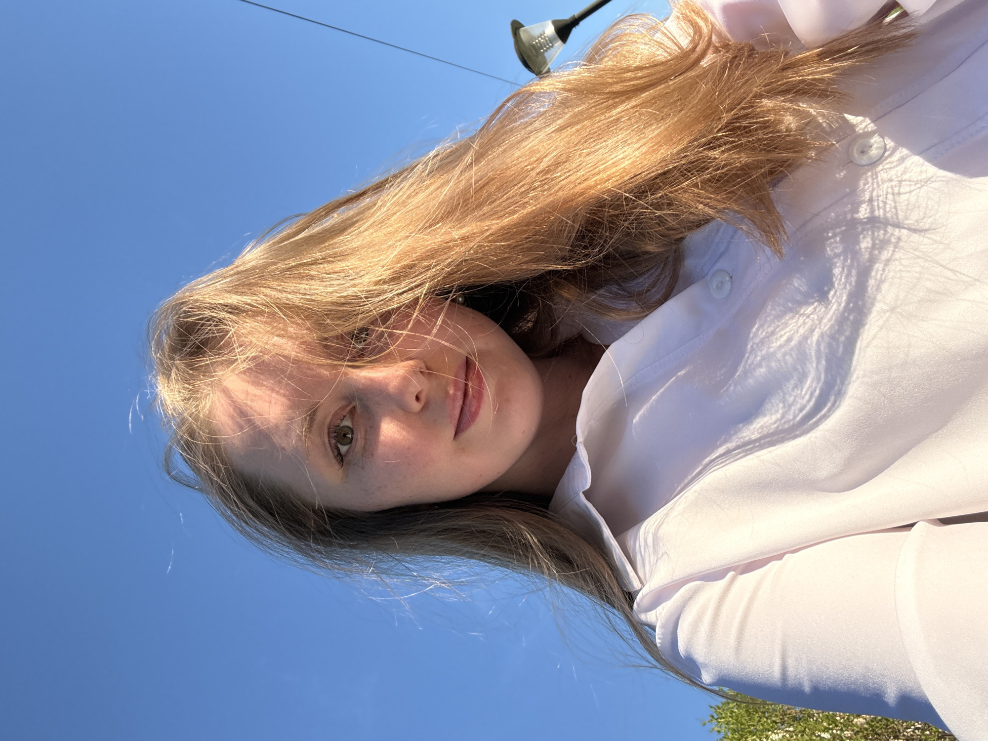 A young, fair-skinned woman is looking down at the camera. She has strawberry blond hair, and is wearing a white button-down shirt. The background is a clear blue sky, and there is a streetlight visible in the distance. The location is Unknown, Unknown.