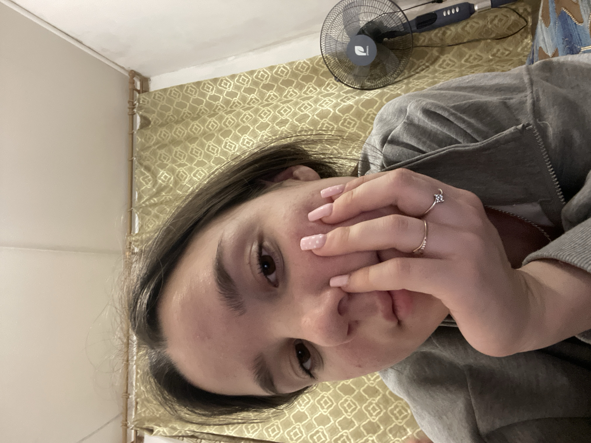 A close-up, angled shot depicts a person with brown hair wearing a gray hooded sweatshirt. Their face is partially obscured by their hand. Two rings are visible on their fingers. In the background are patterned gold curtains and a standing fan with a blue and gray base. The location is Ольховка, Russia.