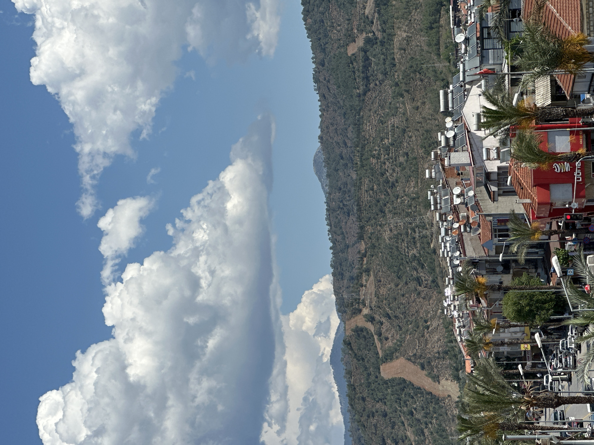 View of Fethiye, Turkey. A line of buildings runs parallel to the mountain. Most buildings are topped with water-heating systems and satellite dishes. Palm trees line the streets on the right. A clear blue sky with white clouds fills the left.