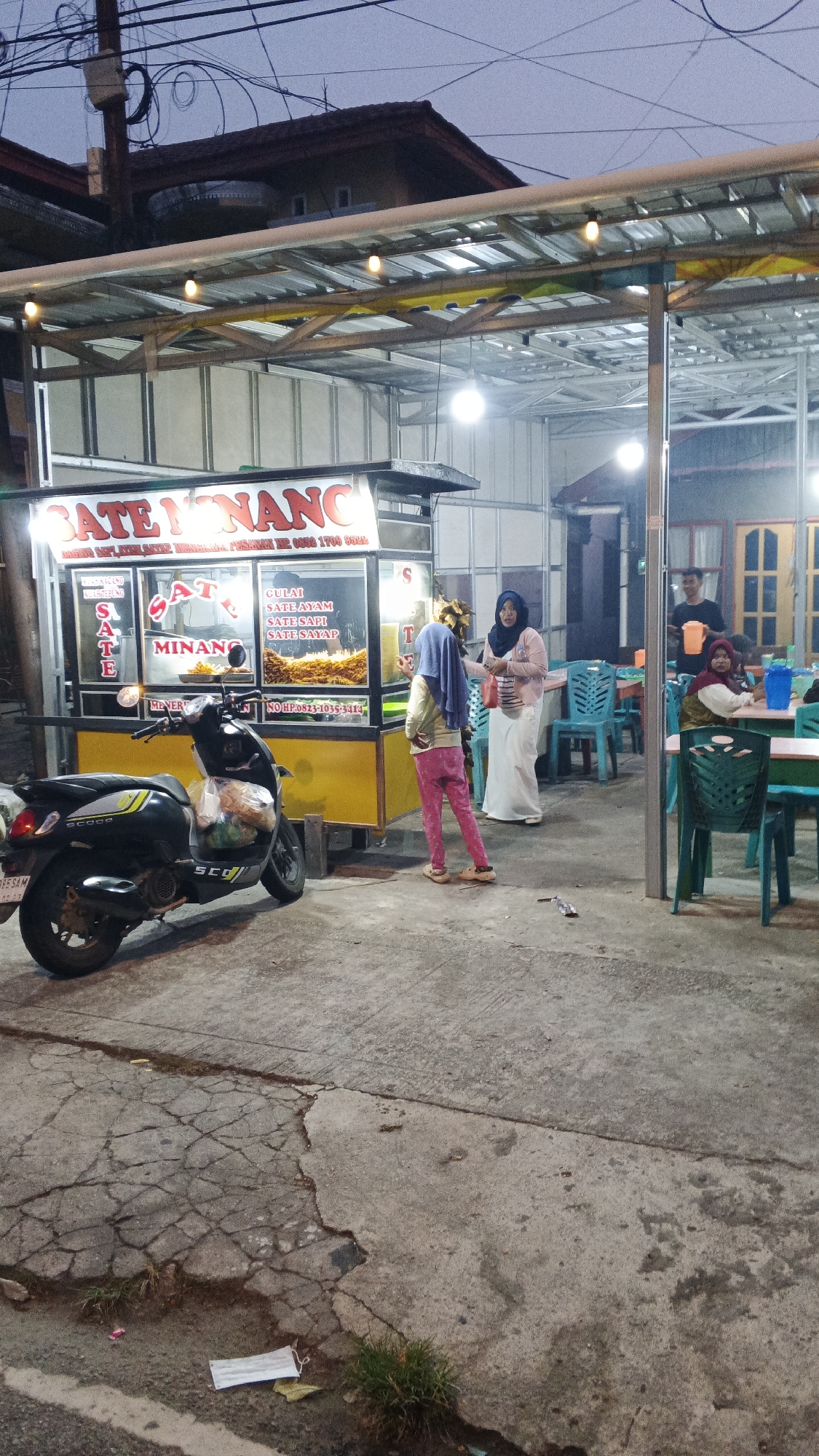 A street-side food stall named "SATE MINANG" is illuminated by string lights under a metal awning. The stall displays menus for various satay dishes like "SATE AYAM" and "SATE SAPI." A black scooter is parked to the left of the stall. Two women, one wearing a pink hijab and a long white skirt, and another, a young girl wearing a blue hijab and pink pants, are standing in front of the stall. In the background, a man is serving drinks at a table, and a woman is seated at another table. The ground is concrete with some cracks and patches of grass. The sky is dark, indicating it's either early morning or evening. The scene appears to be in a commercial area of Siak Sri Indrapura, Indonesia.