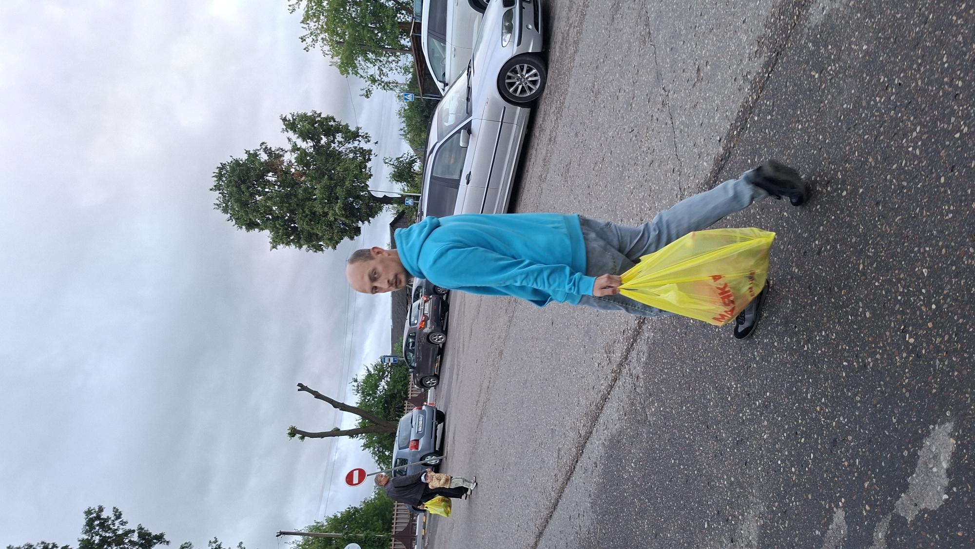 A man wearing a bright blue hoodie and light blue jeans walks on an asphalt road. He is carrying a bright yellow plastic bag filled with some items in his right hand. His left leg is bent at the knee as he walks, and his left arm is extended behind him for balance. He is looking towards the camera with a neutral expression.
In the background, several cars are parked on a paved area. Some trees are visible on the left side of the image, and the sky is overcast, suggesting a cloudy day. The scene appears to be a parking lot or an area near a road, possibly in a town or a suburban area. The text "МАГАЗИН" is visible on the yellow bag. The location is identified as Smolayavichy, Belarus.