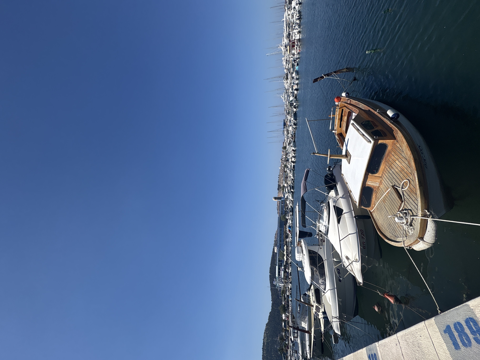 The image shows a marina in the city of Bar, Montenegro, on a clear, sunny day. Several boats are docked, including a small wooden boat in the foreground with a white cabin. Behind it, larger white motorboats are moored. In the background, a vast array of sailboats and motor yachts fill the harbor, with a city skyline and a hill visible in the distance. The water is a deep blue, and the sky is a cloudless azure. The scene captures a typical day in a bustling Mediterranean port.