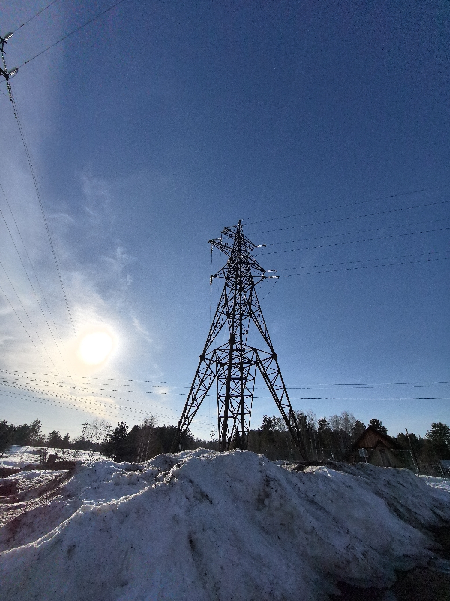 The image shows a high-voltage power transmission tower located in Noviy, Russia. The tower is silhouetted against a bright blue sky with some scattered clouds. The sun is visible in the background, obscured by clouds. The foreground is dominated by large piles of snow. Behind the snow, there are trees and a small building with a gabled roof. Power lines extend from the tower across the sky.