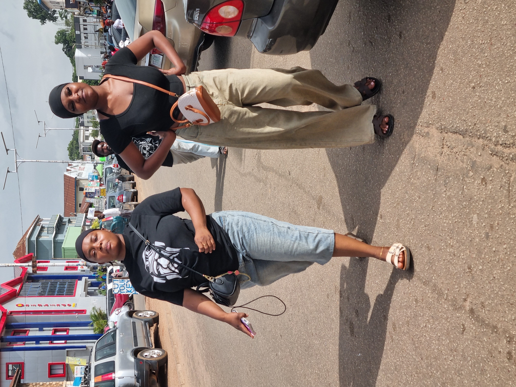 Two women are walking on a paved street in Jos, Nigeria. The woman on the left wears a black t-shirt with a graphic print and light blue denim capri pants, carrying a black shoulder bag. She is holding a cell phone in her right hand. The woman on the right wears a black crop top and loose-fitting beige pants, with a small tan and white crossbody bag. Both women are wearing sandals. In the background, several cars are parked and a building with colorful accents is visible. The sky is overcast.