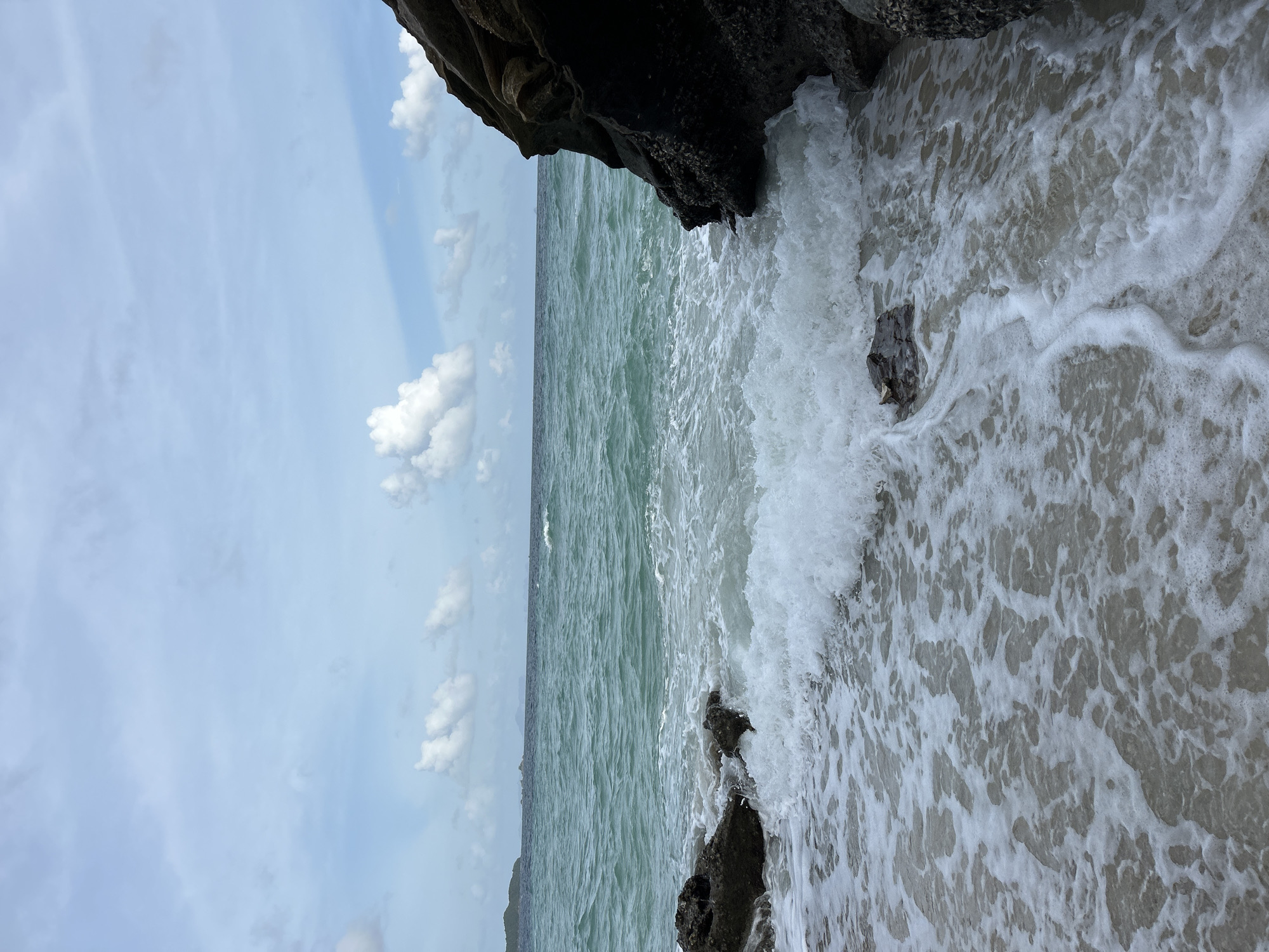 The image depicts a coastal scene in Phru Nai, Thailand. The foreground features the ocean's edge with gentle waves lapping against a sandy shore dotted with rocks. A large, dark rock formation juts out from the right side of the frame, partially submerged by the incoming tide. The water is a clear turquoise color, with white foam and bubbles from the waves. In the distance, the ocean stretches to the horizon, meeting a sky filled with scattered white clouds against a pale blue backdrop. A faint outline of islands or landmasses can be seen on the left horizon. No people are present in the image.