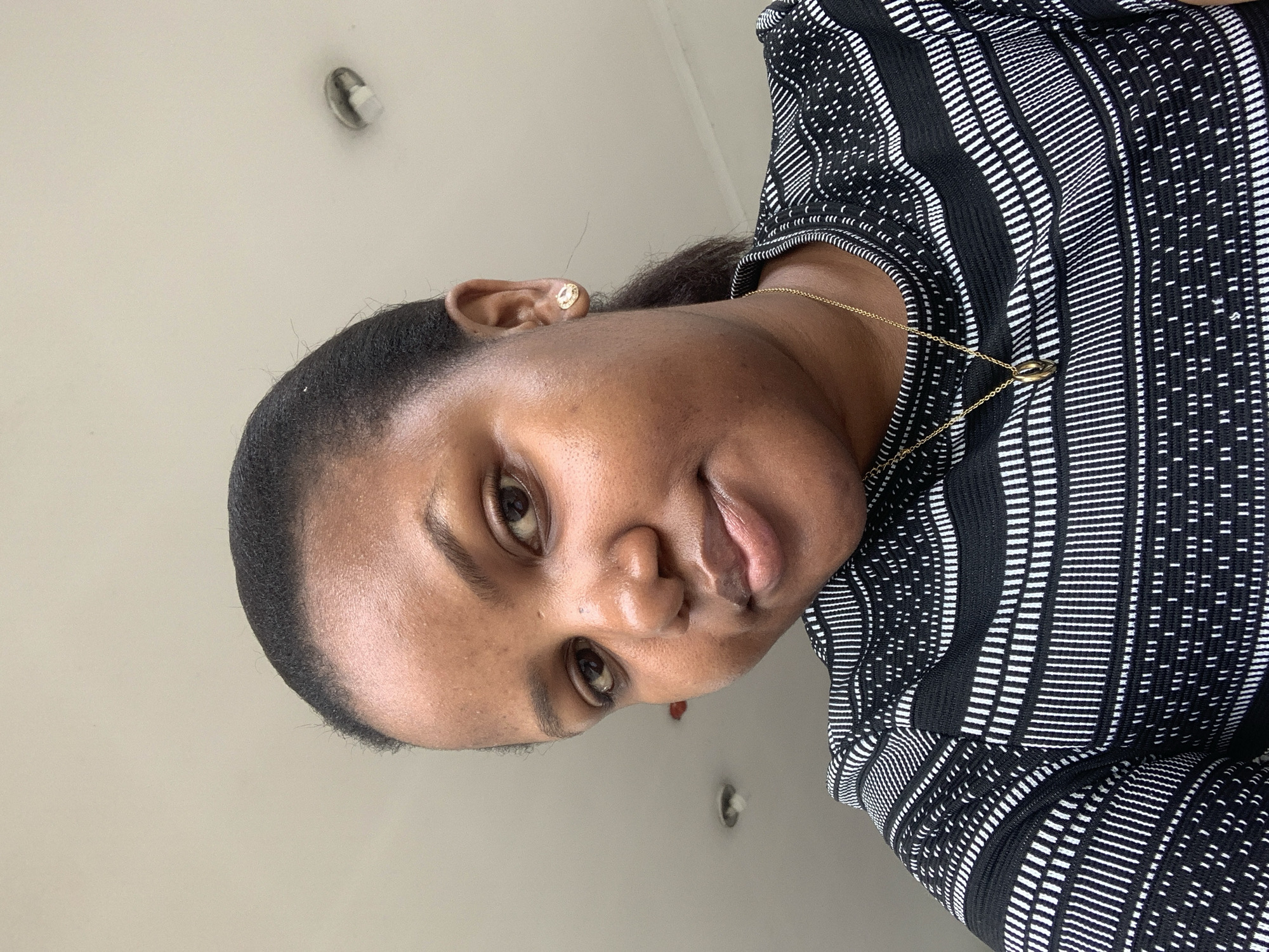 A medium close-up selfie of a young Black woman with dark hair pulled back in a ponytail is shown. She is wearing a black and white horizontally striped shirt and a gold necklace with a pendant. She has on small gold stud earrings. The background is a plain, off-white ceiling with two circular light fixtures. There is no indication of her location in Dar es Salaam, Tanzania.