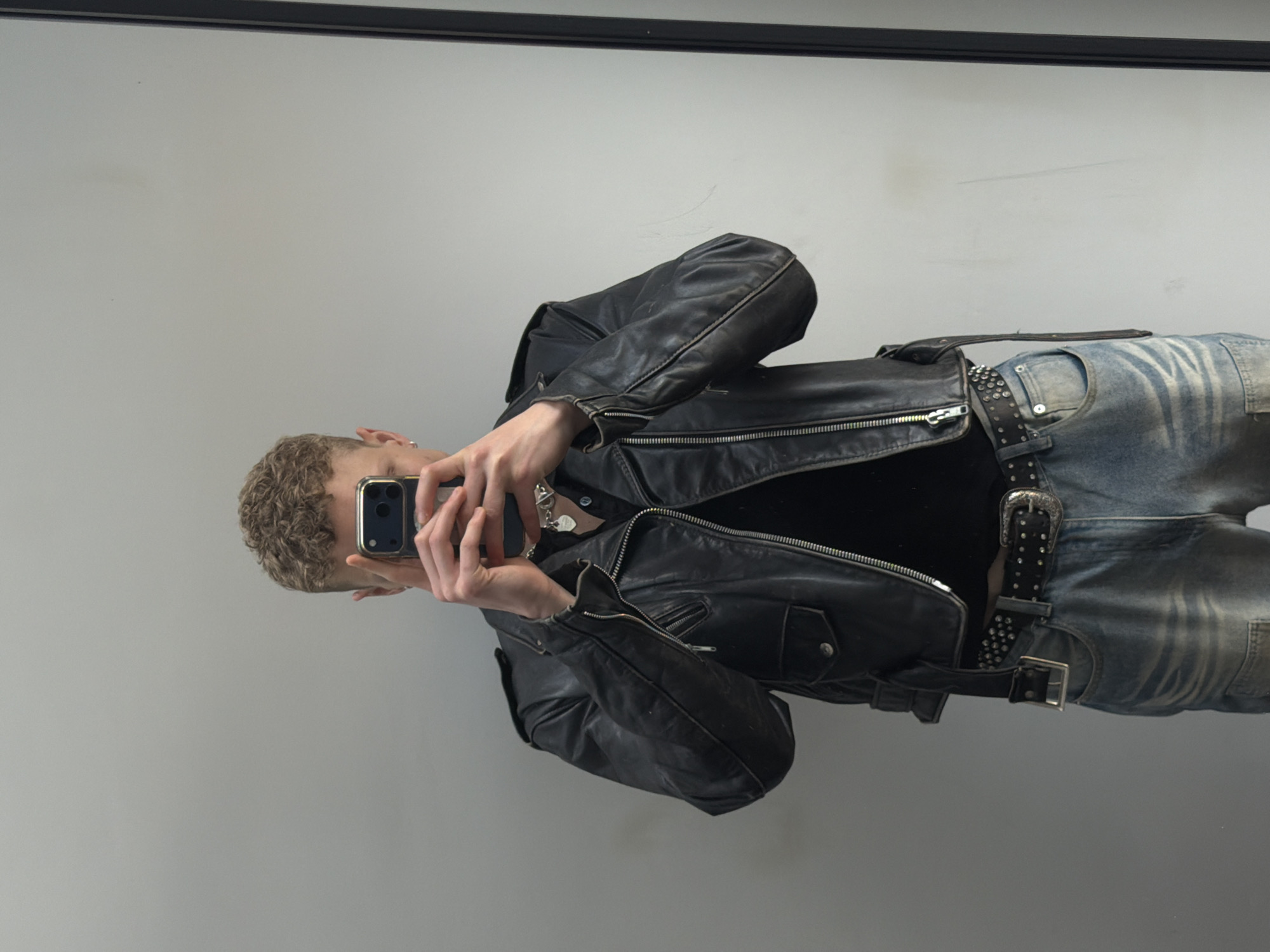 A person with curly blonde hair is taking a mirror selfie with a smartphone. They are wearing a black leather jacket, a black shirt, distressed blue jeans, and a studded belt with a large buckle. A silver necklace is visible around their neck. The background is a plain grey wall with a dark horizontal band at the top, likely part of the mirror frame. The location is not identifiable as London based on the provided information.