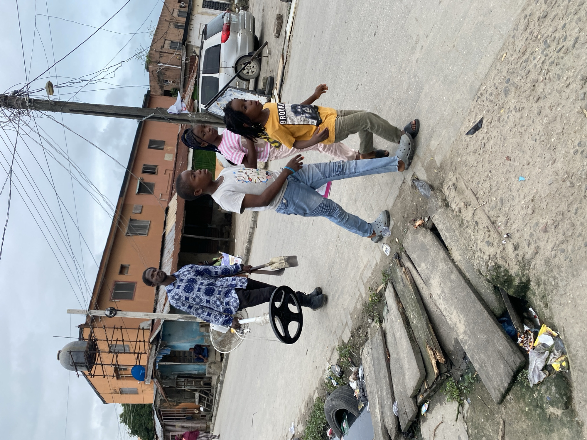 In a city street scene, an adult male walks away from the camera carrying a fan in his left hand and a shovel in his right. He is wearing a blue and white patterned shirt and black pants. Three children walk ahead of him. The foremost child, a boy, is wearing a grey t-shirt and blue jeans. Behind him, a girl in a pink and white striped shirt and light-colored pants walks next to a boy in a yellow t-shirt and green pants. The street is paved and appears to be in a state of disrepair, with debris and refuse visible along the roadside and in a trench. In the background, there are buildings with orange facades and corrugated metal roofs. A silver minivan is parked on the right side of the street. Overhead, a complex network of electrical wires is strung between poles.