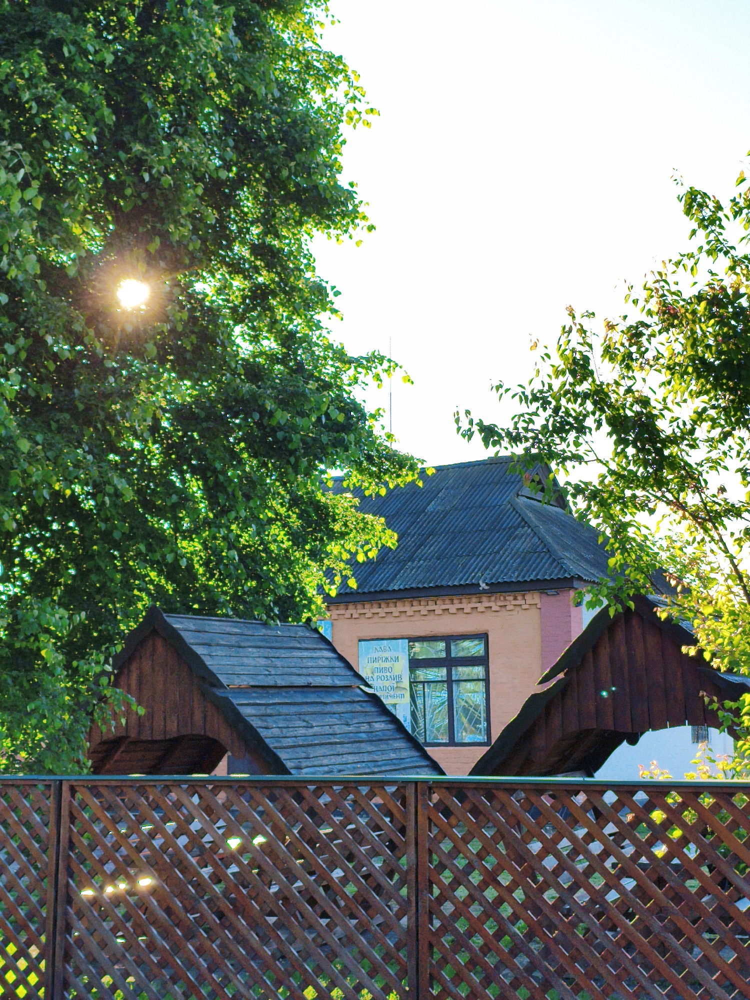 A low-angle shot shows a portion of a building behind a fence in Ustyvytsia, Ukraine. The fence is made of brown lattice wood. Behind the fence, a brown wooden structure sits in front of a two-story building. The building has a grey tiled roof and a light-pink painted facade. A sign is visible on the facade, with writing in a language other than English. Green trees are visible to the left and right of the building. Sunlight shines through the trees. The sky is a pale blue.