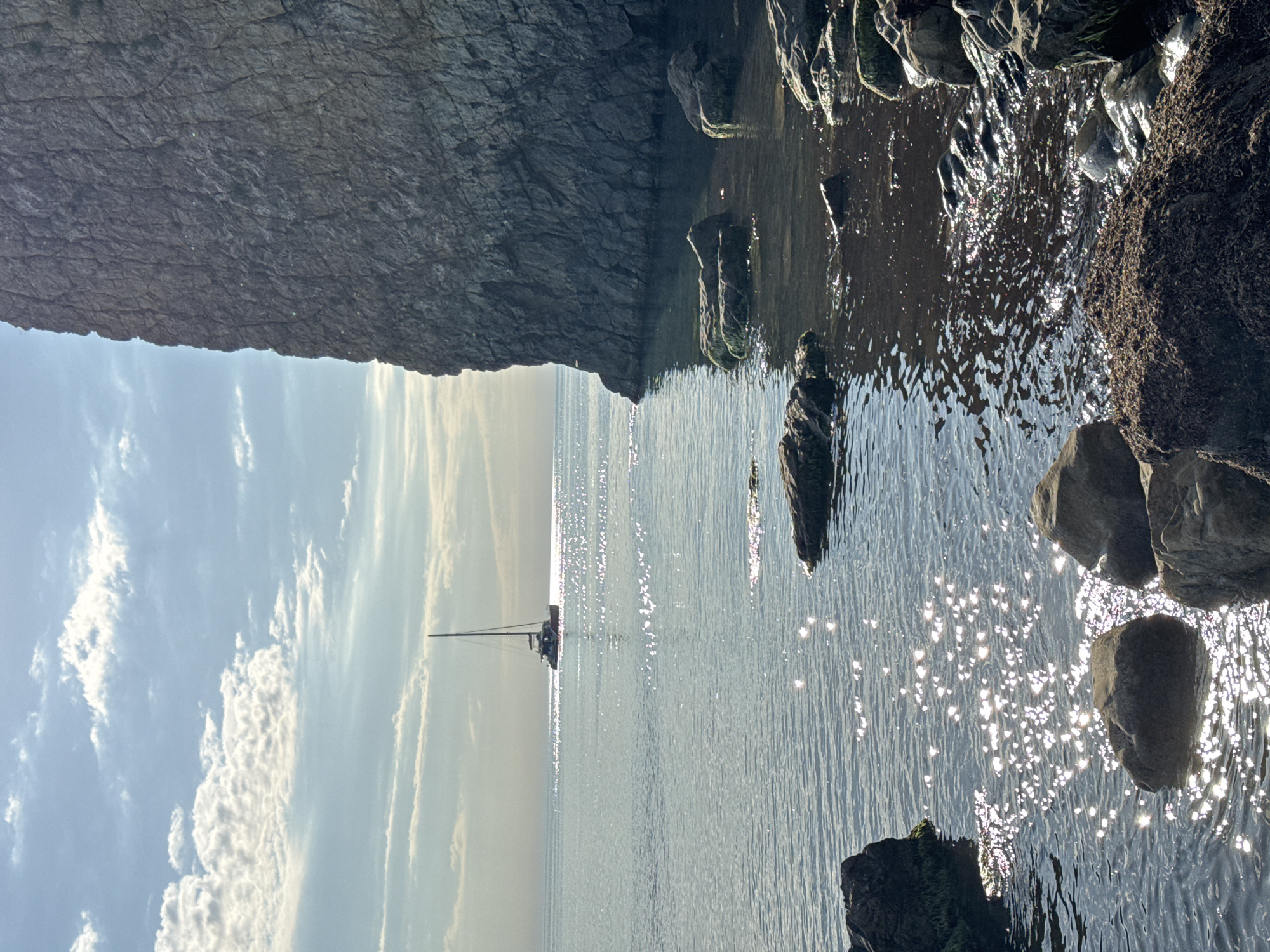 This landscape-oriented image captures a serene coastal scene, likely viewed from within a cave entrance or beneath a large rock overhang. On the left, a massive, dark grey rock formation with intricate, textured surfaces dominates the upper portion, suggesting a cliff face or cave ceiling. Its shadow extends over the tranquil water immediately adjacent to it.
The calm sea stretches towards the horizon, reflecting a dazzling array of sunlit sparkles, particularly towards the right side of the frame. In the foreground on the right, several rugged rocks, some appearing partially submerged and possibly covered in dark moss or algae, break the water's surface. Further out on the calm water, a solitary sailing boat or yacht is visible in the distance, silhouetted against the bright horizon line.
Above the horizon, the sky is a soft blue, adorned with scattered white clouds, particularly noticeable on the upper left. The overall lighting suggests it is daytime, possibly morning or late afternoon, with the sun casting bright glints on the water's surface. No visible text or people are present, emphasizing the natural tranquility of the scene in Divnomorskoe, Russia.