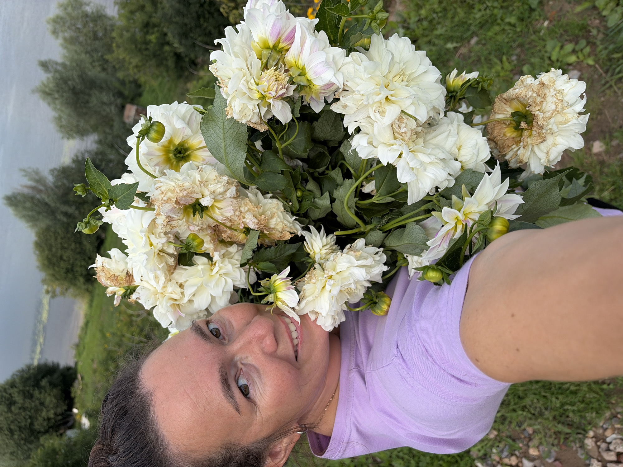 A medium shot captures a woman holding a bouquet of white dahlias in a selfie-style photograph. The woman is positioned on the left side of the frame, with her face turned towards the camera and a smile on her lips. She is wearing a light purple t-shirt. The bouquet of flowers dominates the right and center of the image, with white petals and some browning and wilting edges on certain blooms. The background suggests an outdoor setting, with green grass and trees visible, and a body of water in the distance, possibly a lake or river. The overall scene appears to be in a rural or suburban area, with the natural environment being a prominent feature. The location is identified as Olkhovka, Russia.