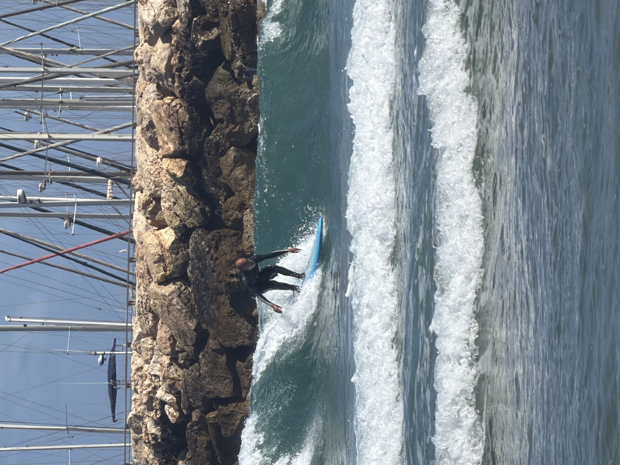 The image shows a surfer riding a wave along a rocky breakwater. The water is choppy with white foam cresting on the waves. The surfer is wearing a dark wetsuit and is standing on a blue surfboard. A marina with masts is visible to the left of the breakwater. This scene likely occurs near a breakwater in Tel Aviv, Israel.