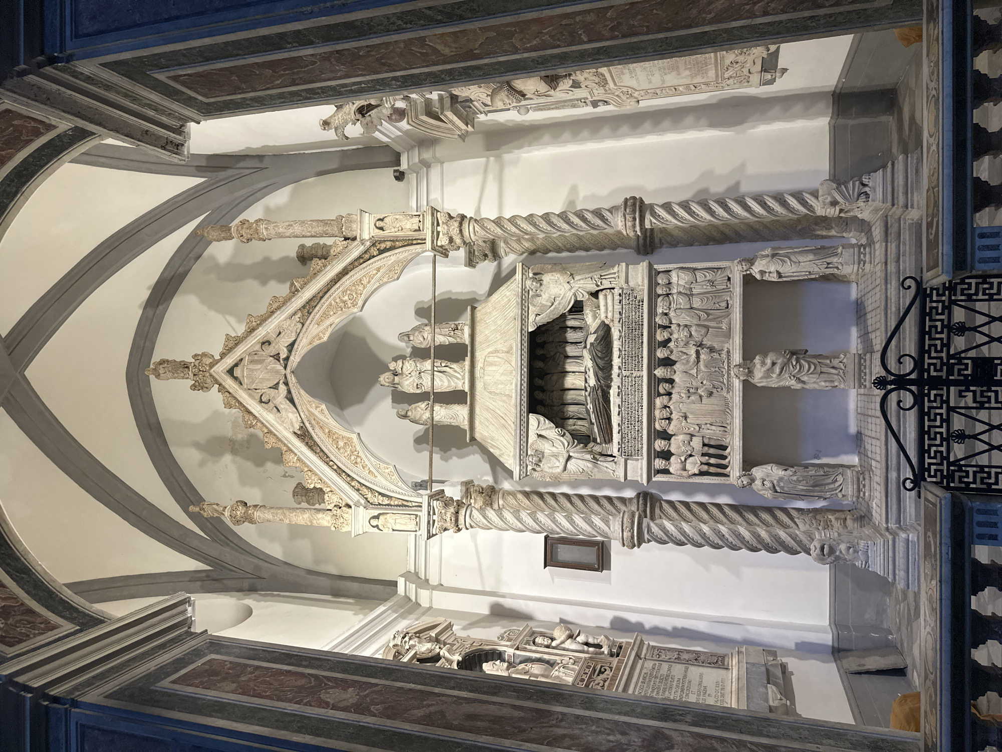 The image shows a detailed view of the San Domenico Maggiore tomb in Naples, Italy. The tomb is an elaborate monument, likely made of marble, featuring numerous sculptures and architectural elements. A sarcophagus rests on a platform, adorned with figures depicting a deceased person lying down, covered by a draped canopy. Above the sarcophagus, a sculptural group of figures is visible. The tomb is flanked by twisted columns and supported by a base with reliefs of standing figures. Intricate carvings and decorative details are present throughout the monument. The background shows a church interior with arched ceilings and decorative walls. The overall scene conveys a sense of historical significance and artistic craftsmanship.