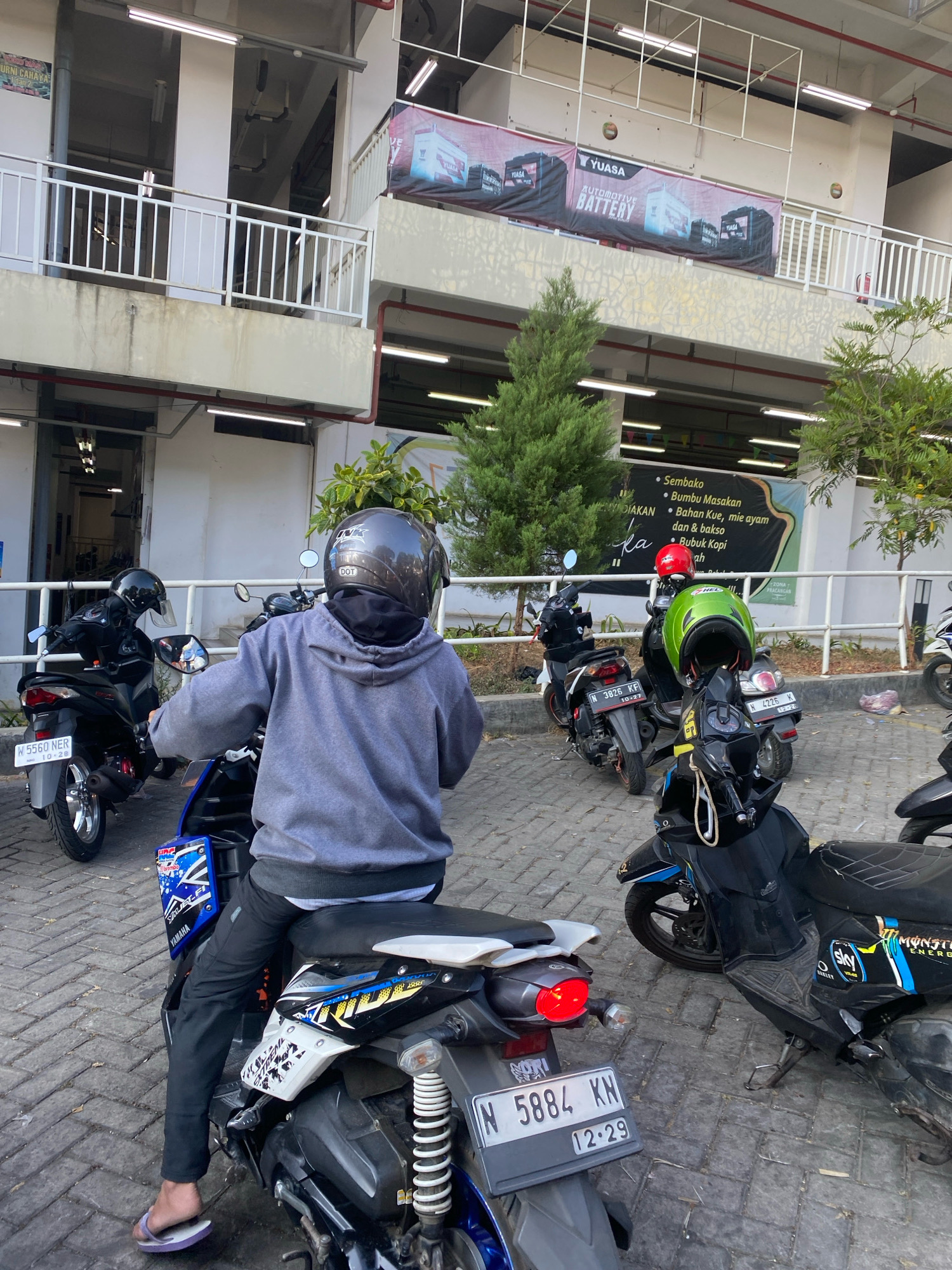 A medium shot shows a person on a scooter parked on a paved surface. The person, viewed from behind, wears a gray hooded sweatshirt, black pants, and flip-flops. They have a dark helmet with a reflective visor and a black scarf partially covering their face. The scooter is dark colored with blue and white decals, and has a license plate that reads "N 5884 KN".
Surrounding the scooter are other parked scooters, some with helmets on them. In the background, there is a multi-story building with white railings and banners. One banner displays an advertisement for "YUASA AUTOMOTIVE BATTERY." Another banner lists various food and drink items. Trees and overhead lighting are also visible in the environment. The overall scene suggests a parking area in a commercial or public space.