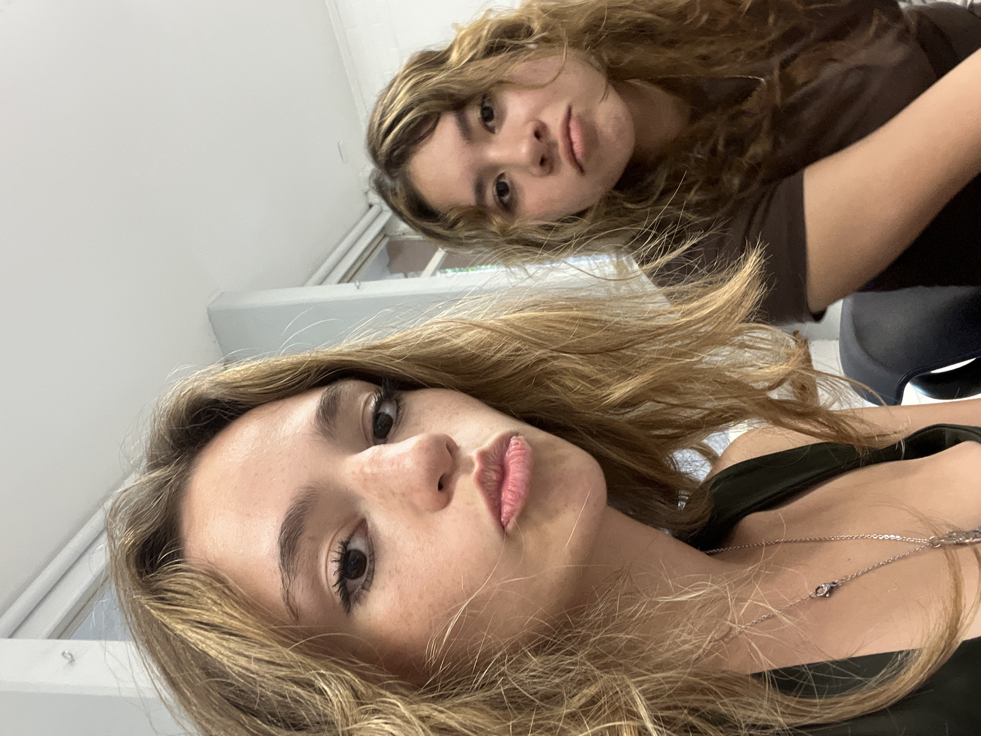 Two young women pose for a selfie in what appears to be an interior setting with white walls. The woman in the foreground has light brown hair, dark eyes, and is wearing a dark top and a silver necklace. Her lips are slightly puckered. The woman in the background has curly brown hair and is wearing a brown shirt. The location is unknown and cannot be determined to be Puebla, Mexico based on the information provided in the image.