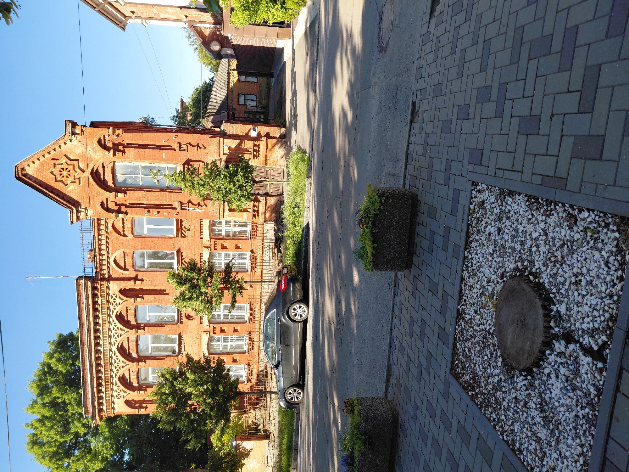 The image captures a sunlit street scene in Poltava, Ukraine. On the left, a prominent, multi-story building constructed from red brick stands with intricate architectural details. It features numerous arched windows with white frames, decorative brickwork including a star-shaped pattern on its gable, and elaborate horizontal bands and cornices. Green trees partially obscure the lower and middle sections of the building's facade.
In the center, an asphalt street runs parallel to the building. A dark-colored sedan is parked alongside the curb, next to a strip of grass. Further down the street, more buildings with tiled roofs are visible.
On the right, a wide sidewalk is paved with grey and darker grey interlocking pavers arranged in a herringbone pattern. Along the sidewalk are several concrete planters containing small green plants. A distinct section of the sidewalk features a bed of white gravel, with a flat, round tree stump embedded in its center.
Bright sunlight casts long, distinct shadows across the street and sidewalk from the trees and buildings, indicating either early morning or late afternoon. The sky above is clear and blue, and no people or discernible text are visible in the tranquil urban setting.
