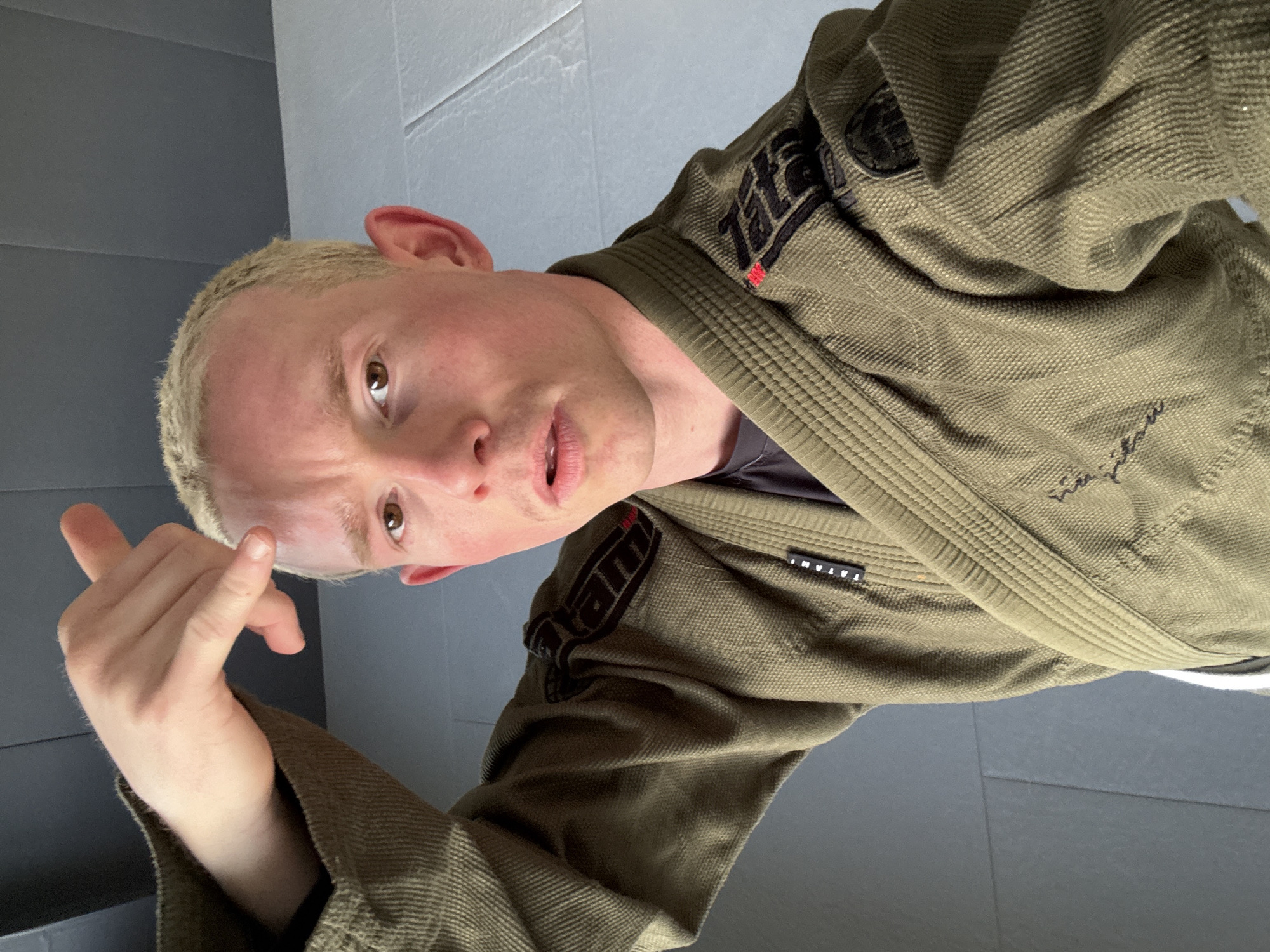 A portrait-style image captures a person, likely male, with fair skin and short, light blonde hair, posing indoors. The individual is wearing an olive green or khaki-colored martial arts gi, characteristic of Brazilian Jiu-Jitsu attire. The gi features a textured weave and black patches with the brand name "TATAMI" visible on both the left shoulder and right lapel. Faint, stylized text, possibly a signature or slogan, can also be seen on the lower right sleeve.
The person is looking directly at the camera with a neutral to slightly inquisitive expression, their brown eyes making direct contact. Their right hand is raised, with the index finger extended and slightly bent, forming a gesture that could imply emphasis, a "come here" signal, or a point of instruction.
The setting appears to be a martial arts studio or dojo, indicated by the dark grey, interlocking floor mats and simple grey walls and a lighter grey ceiling in the background. The lighting suggests an indoor environment, though specific time of day is not discernible. The image has a slightly tilted, dynamic composition, suggesting it may be a selfie. The overall context points to an individual engaged with or presenting aspects related to martial arts training, within the London Borough of Bromley, United Kingdom.