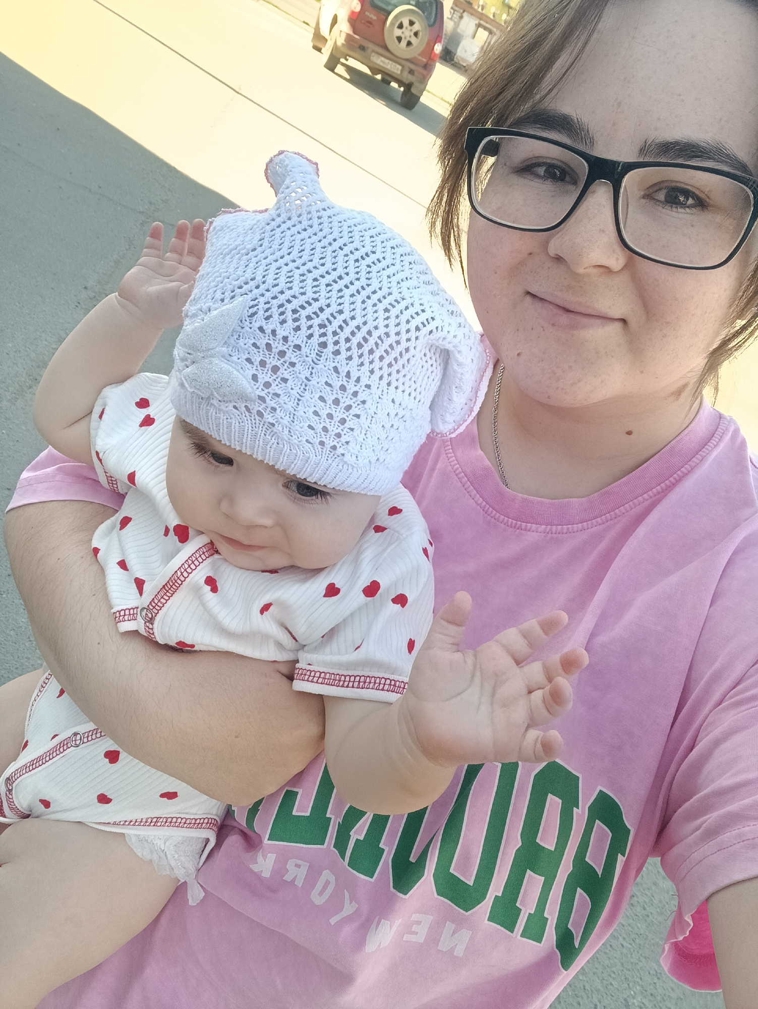 A person with glasses and a pink t-shirt holds a baby in their arms. The baby wears a white patterned bonnet and a white onesie with small red hearts. Both are on a paved surface outdoors. A red car with a spare tire on the back is visible in the background. The location is in the city of Chaikovsky, Russia.