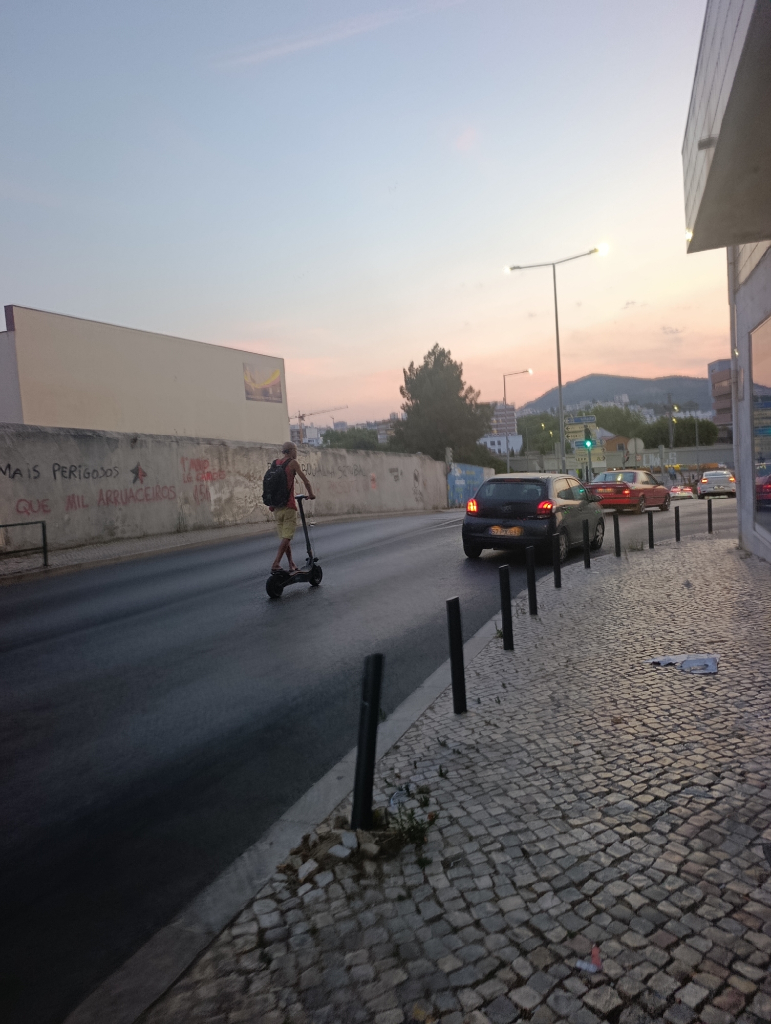 The image depicts a street scene in Setúbal, Portugal, during what appears to be dusk. A person is riding an electric scooter on a wet asphalt road, traveling away from the camera. The rider is wearing a backpack and light-colored shorts. To the left of the road, there is a concrete wall covered in graffiti. In the background, several cars are visible on the road, including a dark hatchback with Portuguese license plates, a red sedan, and a white car. The sidewalk to the right of the road is paved with cobblestones and lined with dark bollards. Beyond the road and cars, there are buildings and a hillside with some greenery. The sky is a gradient of blue and pink, indicating either sunrise or sunset. Streetlights are illuminated.