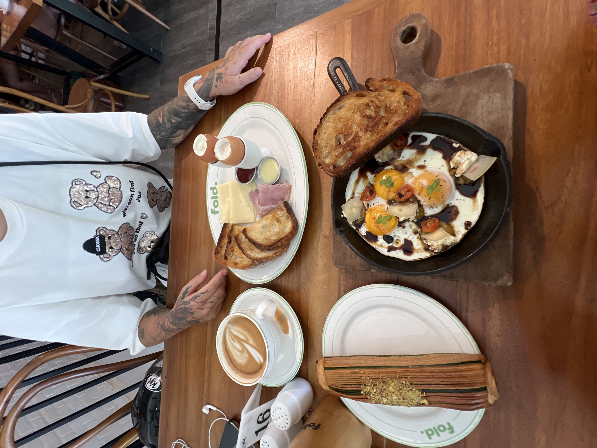 A top-down shot depicts a breakfast scene at "Fold" cafe, presumably located in North Kuta, Indonesia. A light-skinned person with tattooed arms sits to the left of the frame. They are wearing a white shirt with a teddy bear graphic. In front of them on a wooden table are four plates. The plate closest to the person holds boiled eggs, slices of cheese and ham, and toast. To the right, a cast-iron skillet holds fried eggs with a dark sauce, accompanied by a slice of toast leaning against its rim, resting on a wooden serving board. Another plate contains a pastry topped with green sprinkles. A cup of coffee is positioned between the first and third plates. Other items visible on the table include headphones, a number card, and condiments. The setting appears to be indoors, with wicker chairs partially visible in the background.