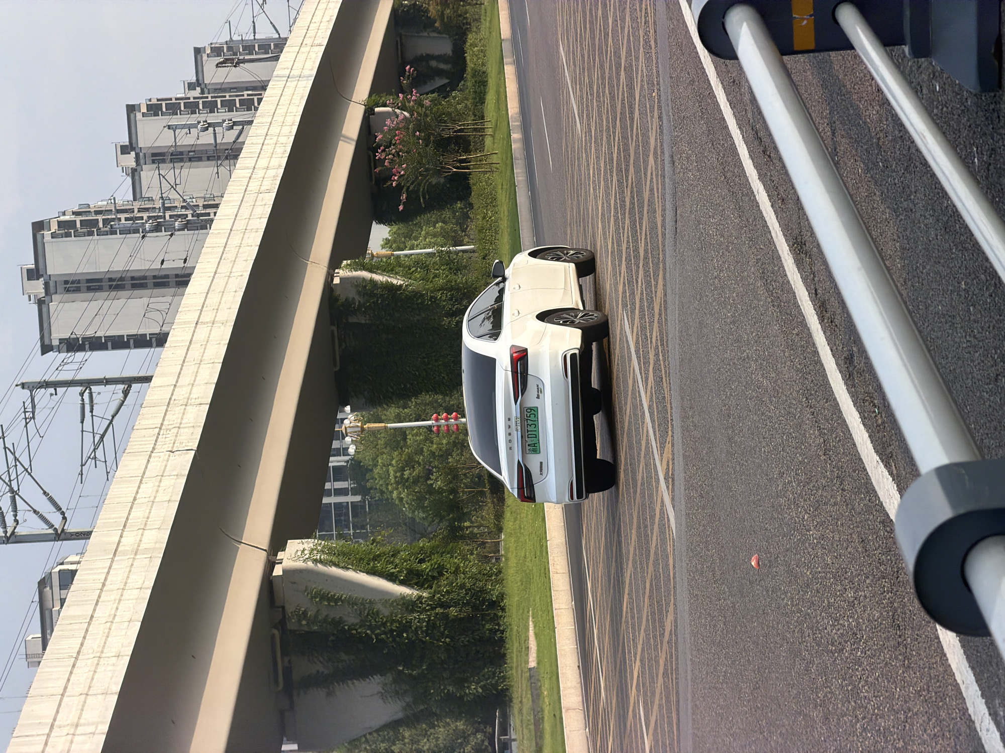 A white sedan with license plate A·DT3759 is captured in motion on a road in Jiangjin, China. The car is positioned in the right lane of a multi-lane road, with its rear facing the viewer. Yellow criss-cross markings on the asphalt indicate a waiting or designated area. In the background, a large, elevated concrete structure, likely an overpass or bridge, dominates the scene. This structure is supported by massive concrete pillars, with greenery growing on and around them. Beyond the overpass, several modern, multi-story buildings are visible, suggesting an urban environment. Overhead, power lines and electrical infrastructure are strung across the sky. The sky is overcast with a light haze. To the right of the road, a lamppost with red decorative elements and trees line the median. In the foreground, a section of a white railing and a grey concrete post are visible, partially obscuring the road. A small, pinkish object is present on the road surface in the middle ground.
