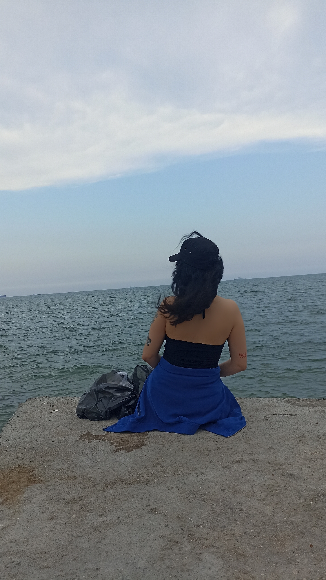 A person wearing a black baseball cap and a strapless black top is sitting on a concrete platform by the sea. A blue cloth is draped over their lap. To the left of the person, there are two black plastic bags. The sea is choppy and extends to the horizon, where a hazy sky with light clouds is visible. Several distant ships are scattered across the sea. The concrete platform appears weathered and shows signs of dampness. The setting is the coast of Yenişehir, Turkey.