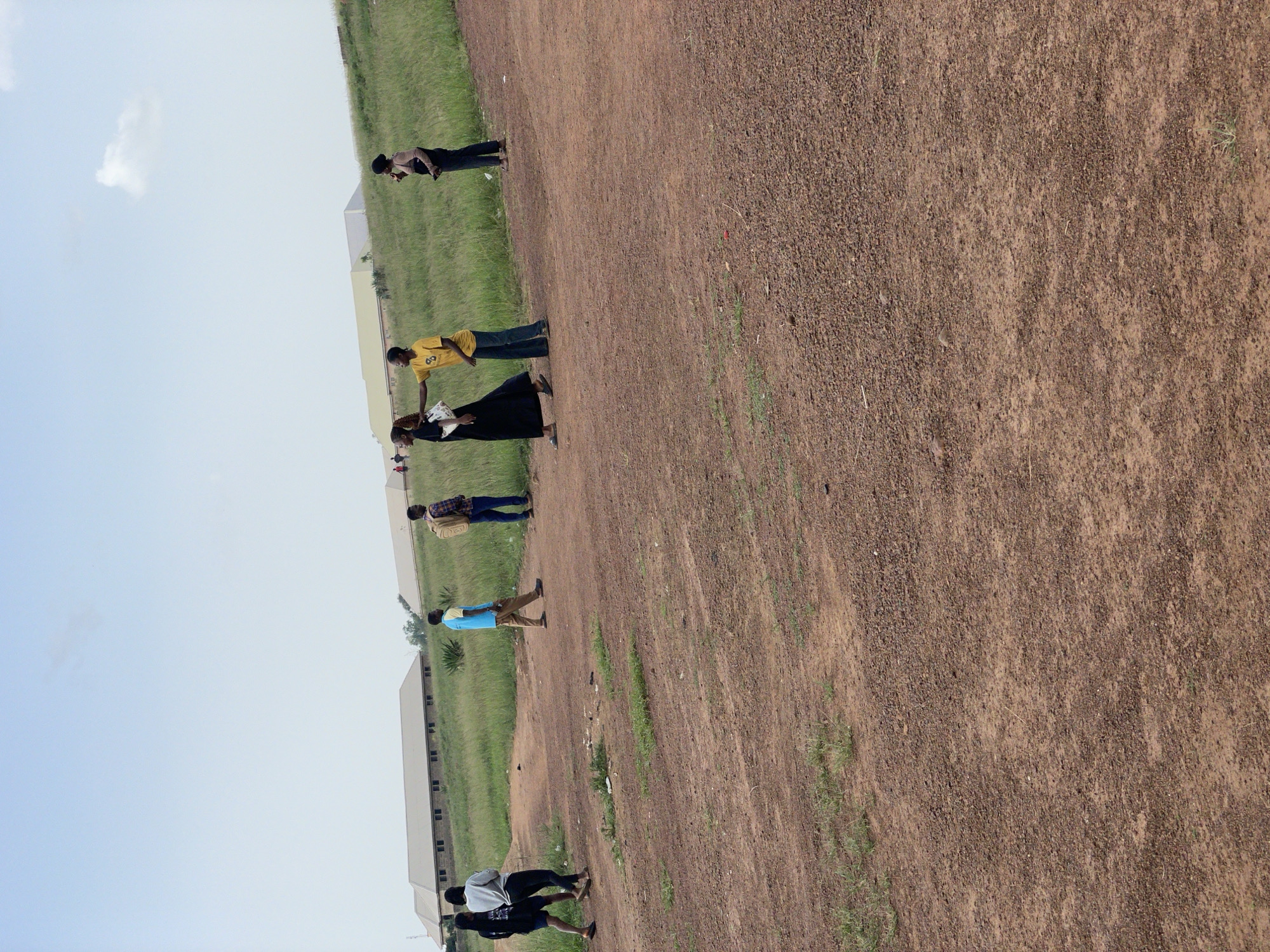 In Durumi, Nigeria, a group of seven individuals are scattered across a dusty, outdoor area. Two women in the foreground, one wearing a black dress and the other a white top, walk together. To their right, another woman in a yellow t-shirt and jeans stands with her hand extended towards the first woman. Further to the right, a man in a patterned shirt and jeans walks away from the viewer, carrying a backpack. To the far right, a person in a dark top and pants stands looking down at a phone. In the background, two more individuals are walking. They are positioned in front of a row of light-colored, rectangular buildings with dark windows, situated on a grassy hill. The sky above is a pale blue with a few wispy clouds.