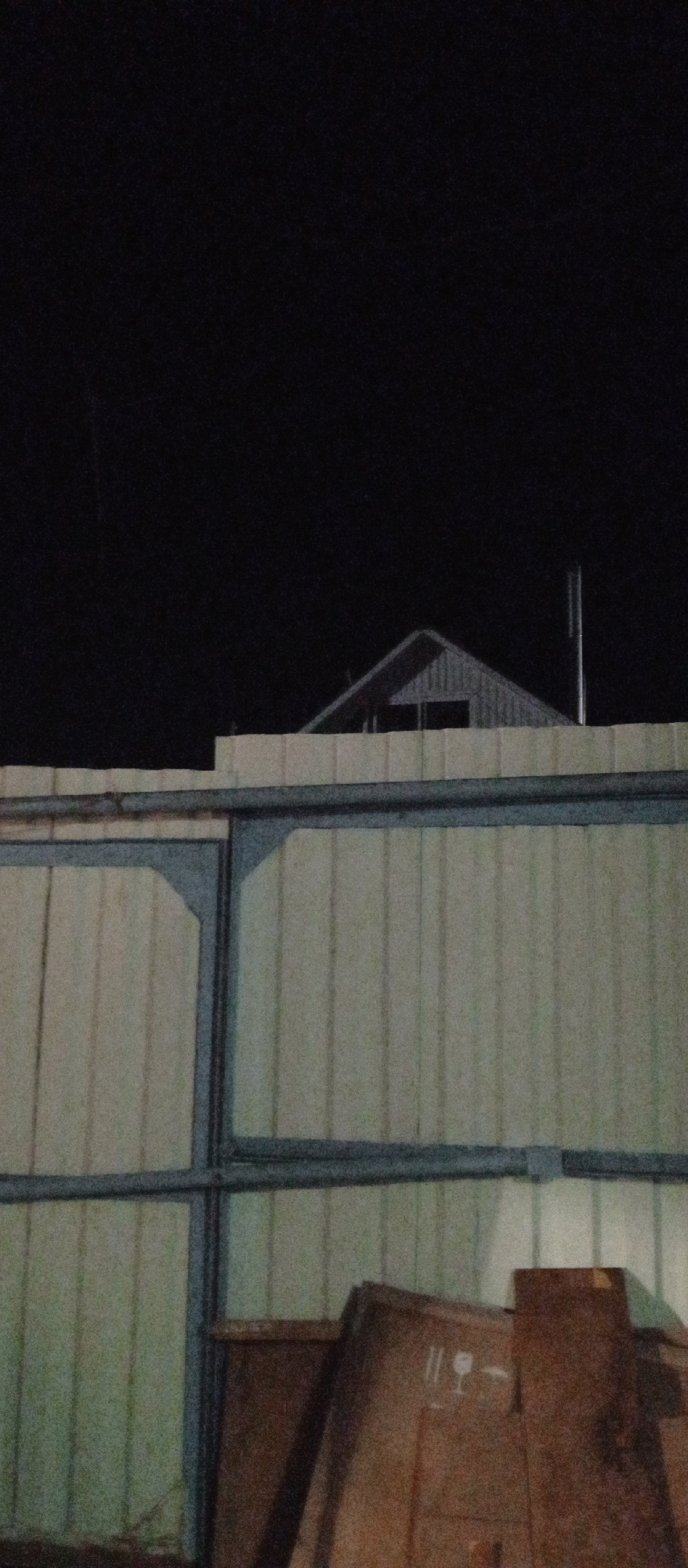 The image shows the roof of a building with a chimney against a dark night sky. In the foreground, there is a corrugated metal wall with horizontal beams. Partially visible at the bottom right is a large wooden crate with markings. The scene appears to be outdoors, possibly in an industrial or construction area, with no people or discernible actions. The location is identified as Nazarovo, Russia.