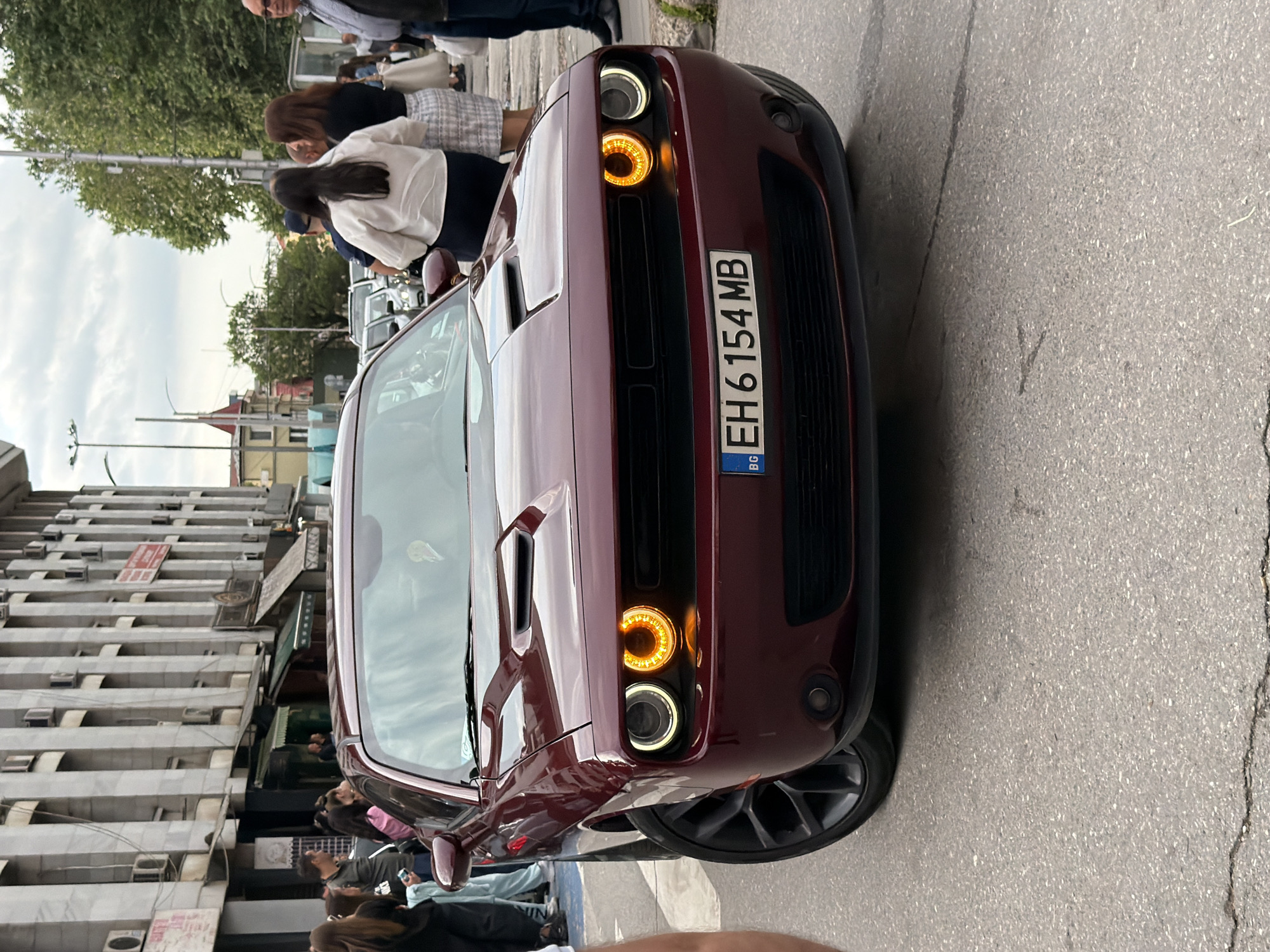 A reddish-brown Dodge Challenger is parked on a street in Pleven, Bulgaria. Its license plate number is EH6154MB. Several people are walking around the car and on the sidewalk in front of a building with numerous windows and signs. The street appears to be busy.
