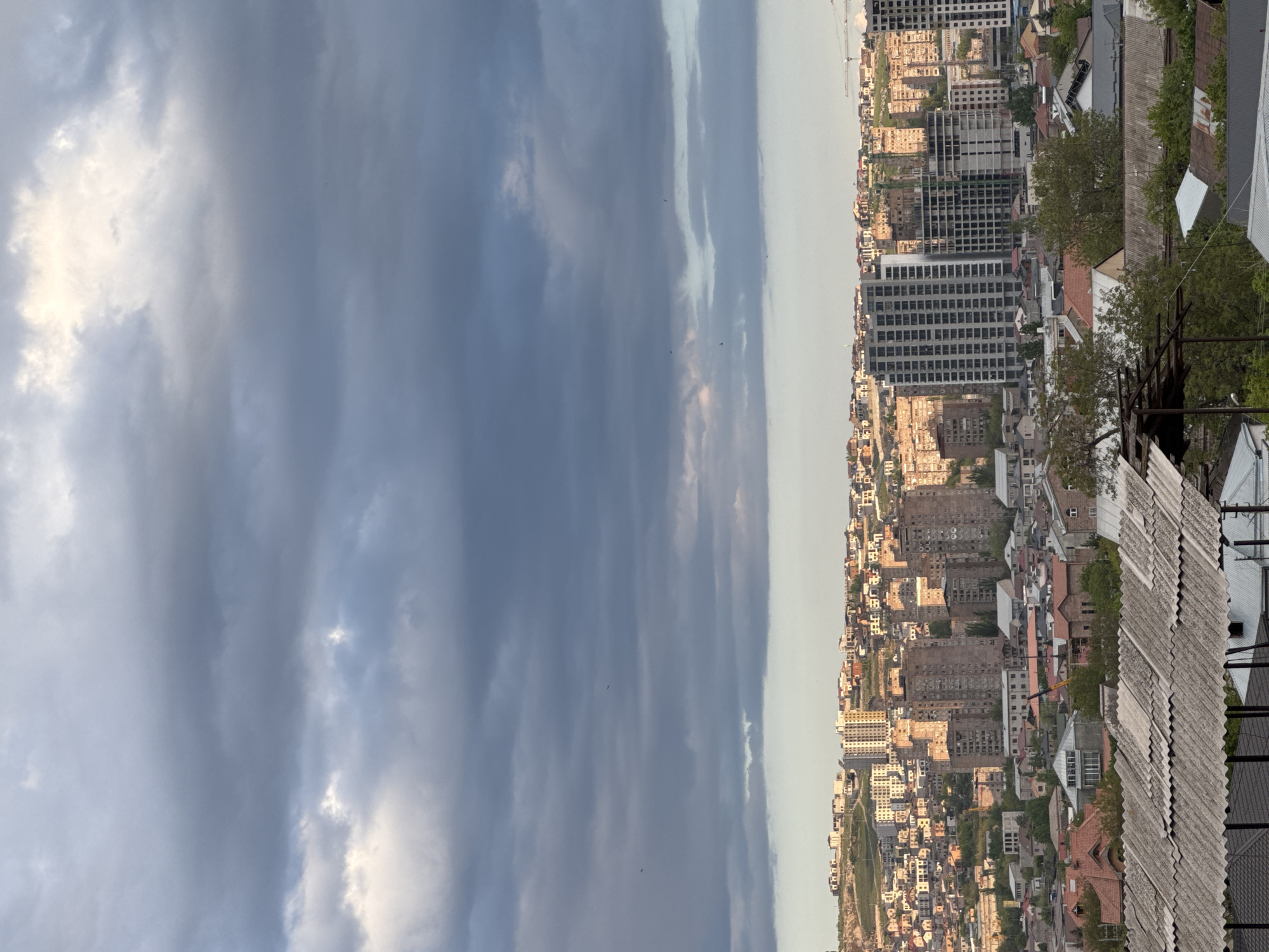 A cityscape scene viewed from an elevated perspective, possibly a rooftop, dominates the right side of the image, while a vast sky filled with dramatic, dark clouds takes up the left half.
The city features an array of buildings, varying in height and architectural style. Some buildings appear to be older with red-tiled roofs, interspersed with more modern, taller structures. A few construction cranes are visible, indicating ongoing development in the area.
The foreground contains parts of the rooftop, including metal railings and roofing materials.
The sky shows a range of grey tones, suggesting a potential storm.
Based on the information available, the location is Arinj, Armenia.