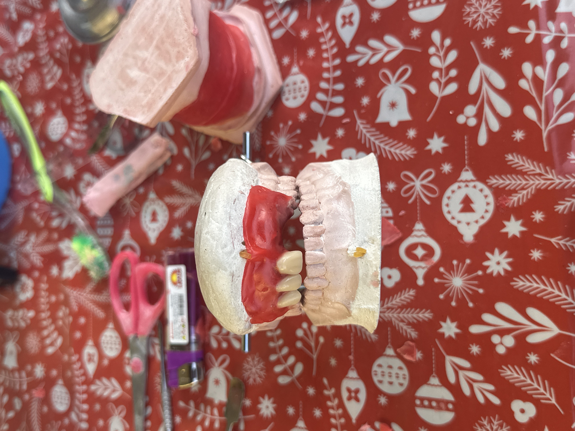 The image shows a dental model composed of two parts, one upper and one lower, mounted on a metal rod and displaying artificial teeth set in red wax. Additional dental models and dental tools, including a pink pair of scissors, are scattered on a red surface featuring a white winter holiday pattern. The location is indeterminate as there are no distinctive features identifying it as being in Parroquia Ocumare, Venezuela. There are no people visible in the image. The objects are dental models and tools, and the primary subject is the dental model in the center of the frame. No actions or interactions are depicted.