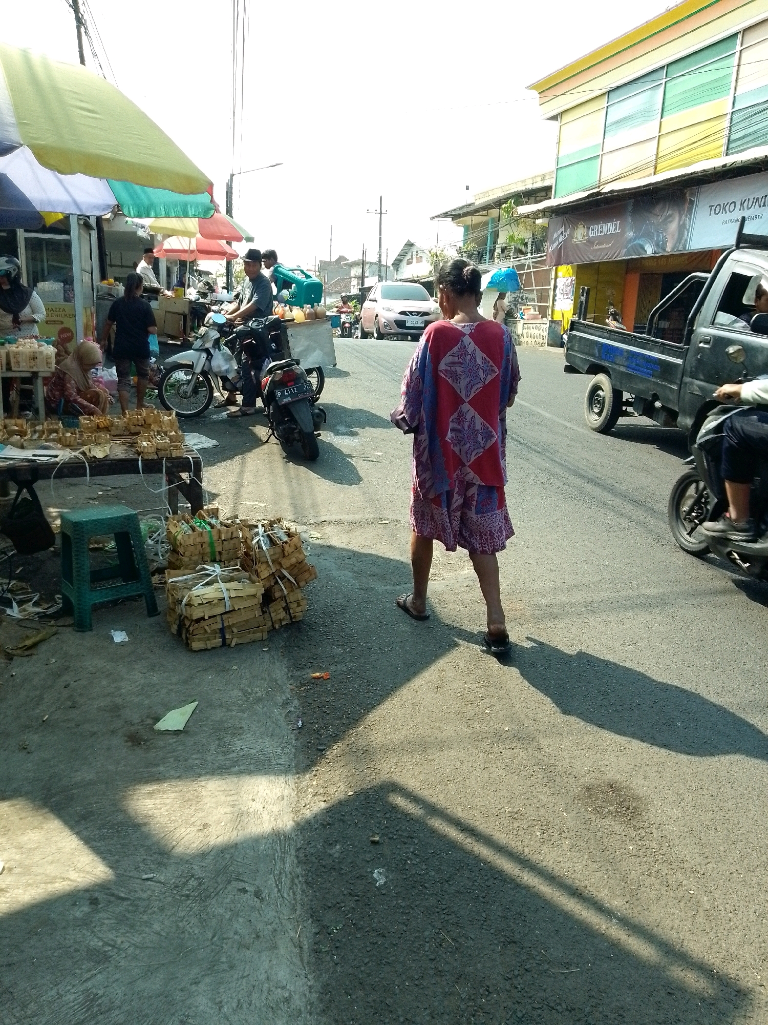 The image depicts a street market scene in Jember, Indonesia. A woman wearing a red and purple patterned garment walks away from the camera. In the background, there are various stalls with umbrellas, motorbikes, and a black pickup truck. A building with colorful windows is visible on the right. The ground is paved with asphalt and concrete, with shadows cast by the sunlight. Several stacks of goods, possibly food items wrapped in straw or paper, are placed on the ground and on a table. A green plastic stool is near the table. The overall impression is a busy outdoor market on a sunny day.