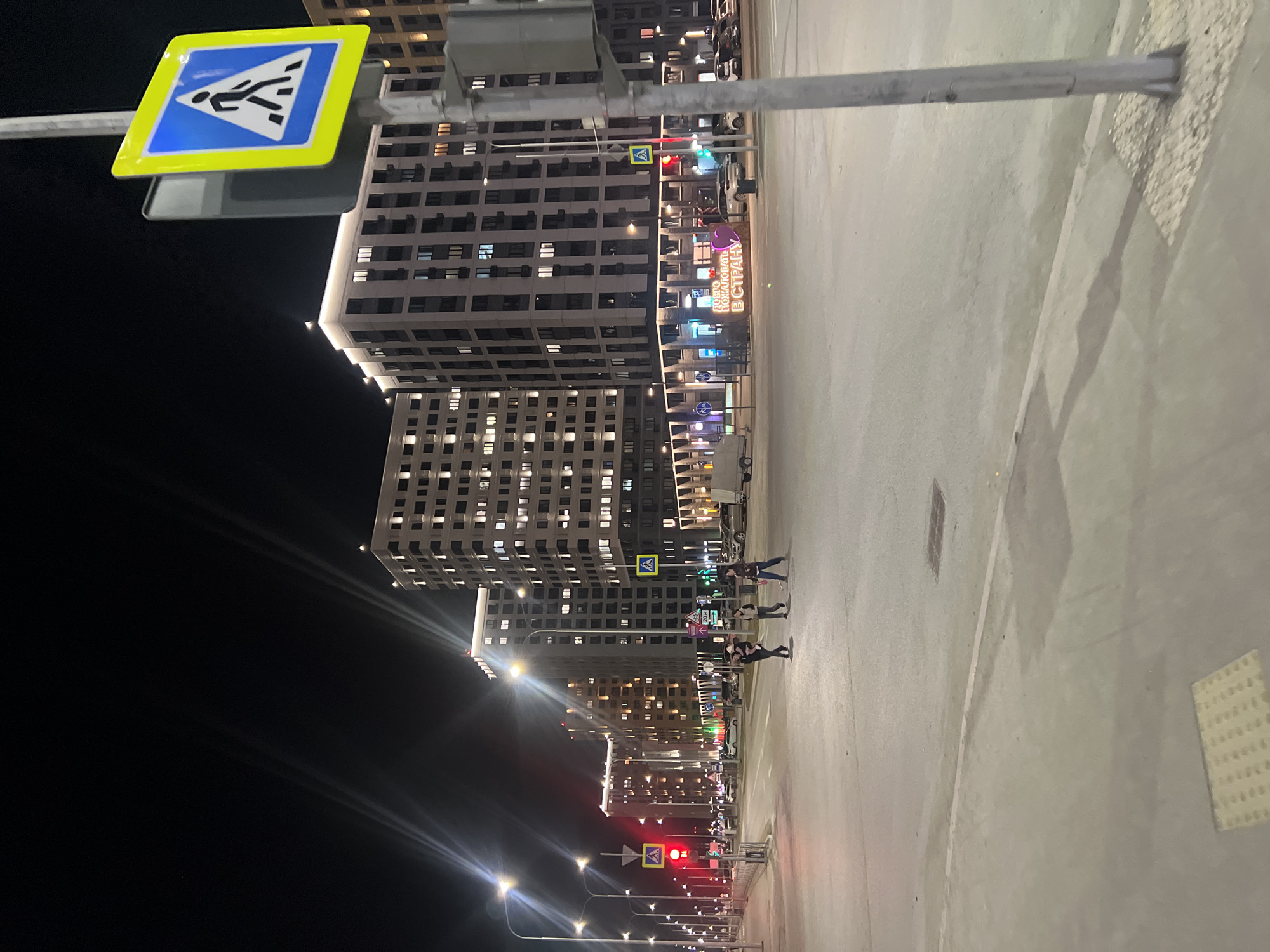 Here is a description of the image:
The image is a nighttime shot taken in what appears to be a modern urban area, likely the city of Tyumen, Russia. Several high-rise apartment buildings are visible in the background, lit up with numerous windows. The buildings have a repetitive, geometric architectural style. A pedestrian crosswalk sign is prominent in the foreground, indicating a safe area for pedestrians to cross the street. The street is wide and paved, showing painted lane markings and some surface details. Streetlights illuminate the scene, casting a bright glow on the road and surrounding area. Pedestrians are visible crossing the street at the crosswalk. Traffic lights are seen in the distance showing red and green signals. The sky is dark and clear.