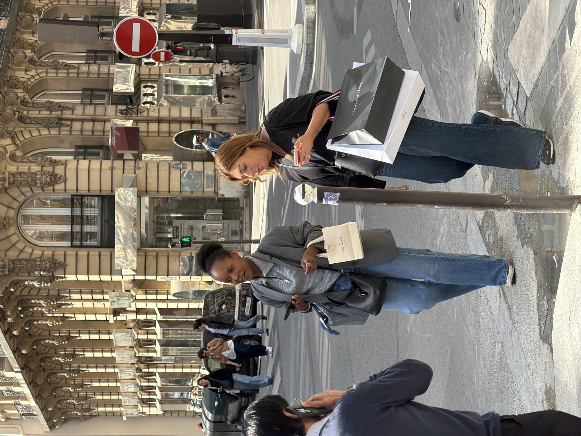 The image shows a street scene in Paris, France. A woman with brown hair holds a shopping bag marked with "Tommy Hilfiger." Another woman with dark hair in a bun holds a small bag marked "Sezane." Both women are wearing jeans. A man with black hair is on the ground, holding a phone up to his ear. Several other people are walking along the sidewalk. Cars and buildings are visible in the background. There are street signs, including a "do not enter" sign. The architecture of the buildings is ornate and typical of Parisian style. The scene is illuminated by sunlight.