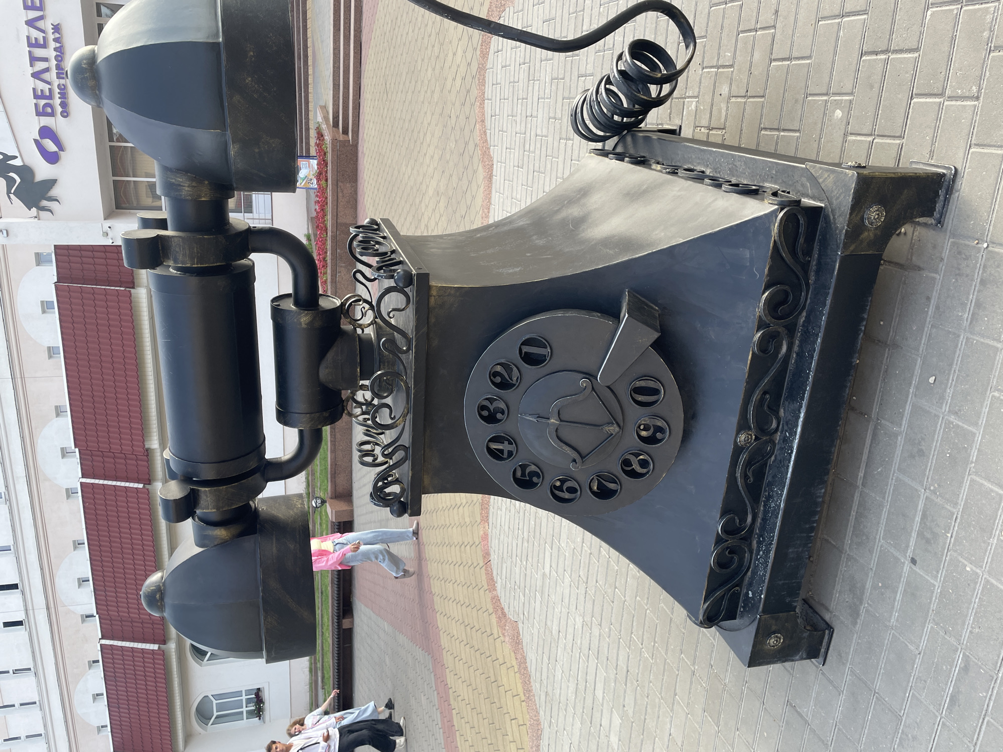 The image shows a large, black, decorative sculpture of an old-fashioned rotary dial telephone. The sculpture is positioned outdoors on a paved walkway. In the background, there are buildings with red tiled roofs and some greenery.
Several people are visible near the sculpture. Two individuals, possibly tourists, are standing and looking at the sculpture. One person in a pink jacket is walking away from the camera, while another person is partially visible in the lower right corner.
The sculpture itself is detailed, with a coiled cord and a dial featuring numbers and a decorative arrow symbol. The metalwork is ornate, with decorative scrollwork along the edges of the base.
The pavement is made of interlocking bricks in shades of beige and pink. The scene appears to be taking place during the daytime, under clear skies, suggesting pleasant weather.
A sign with Cyrillic text is visible on one of the buildings in the background. The text reads "БЕЛТЕЛЕКОМ" and "ОФИС ПРОДАЖ". This indicates a telecommunications office. Given the location context of Brest, Belarus, this is likely a street in that city. The overall mood of the image is casual and public, capturing a moment of everyday life with a unique artistic element.
