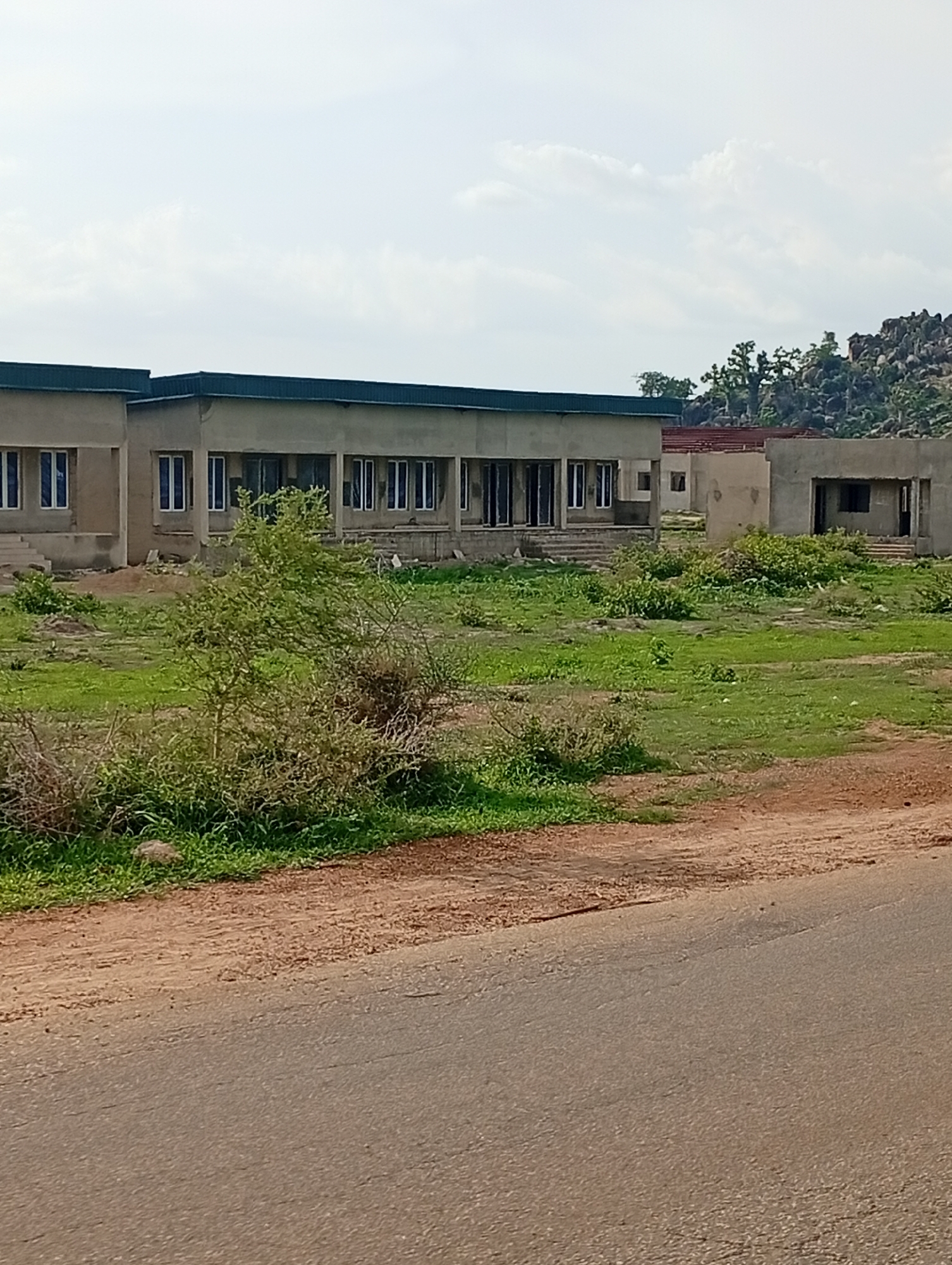 The image shows a row of unfinished concrete buildings under a cloudy sky. The buildings appear to be single-story structures with dark-colored roofs. They have numerous windows, many of which are recessed. The buildings are situated in a grassy, undeveloped area with some scattered bushes and trees. In the background, there are rocky hills with sparse vegetation. A dirt road is visible in the foreground, with an asphalt road bordering it. The scene suggests a construction site or a developing area in a rural or semi-rural environment. The city of Shuwa, Nigeria is mentioned as the location, implying this is a view of buildings and surroundings within or near that area. There are no people visible in the image.