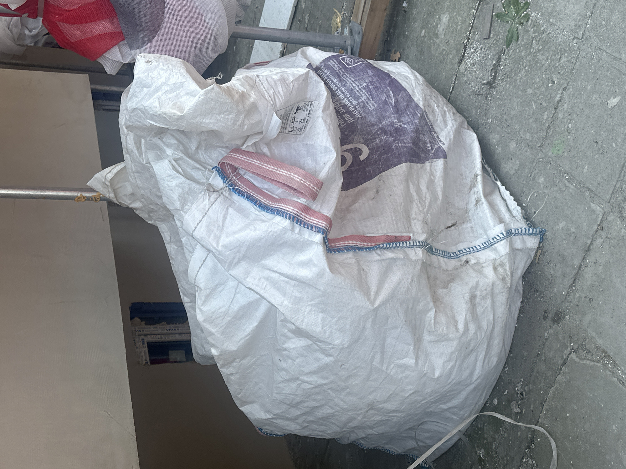 A large, white, woven polypropylene bulk bag lies on a paved surface. The bag is partially filled and slumped, with its opening visible at the top. It has red and blue striped webbing handles and trim along the opening. A label with various symbols and text, including what appears to be "ISO 9001, ISO 22000: 2018", is attached to the bag. A large, purple graphic with white lettering, possibly indicating a product name or brand, is visible on the side of the bag. Some debris, including small stones and a green leafy plant, is scattered on the paved ground around the bag. In the background, a scaffolding pole is visible, and part of a building or wall with what looks like a recessed opening is present. To the right, other bulk bags, one with red and white stripes, are partially visible. The scene suggests an outdoor construction or renovation area.