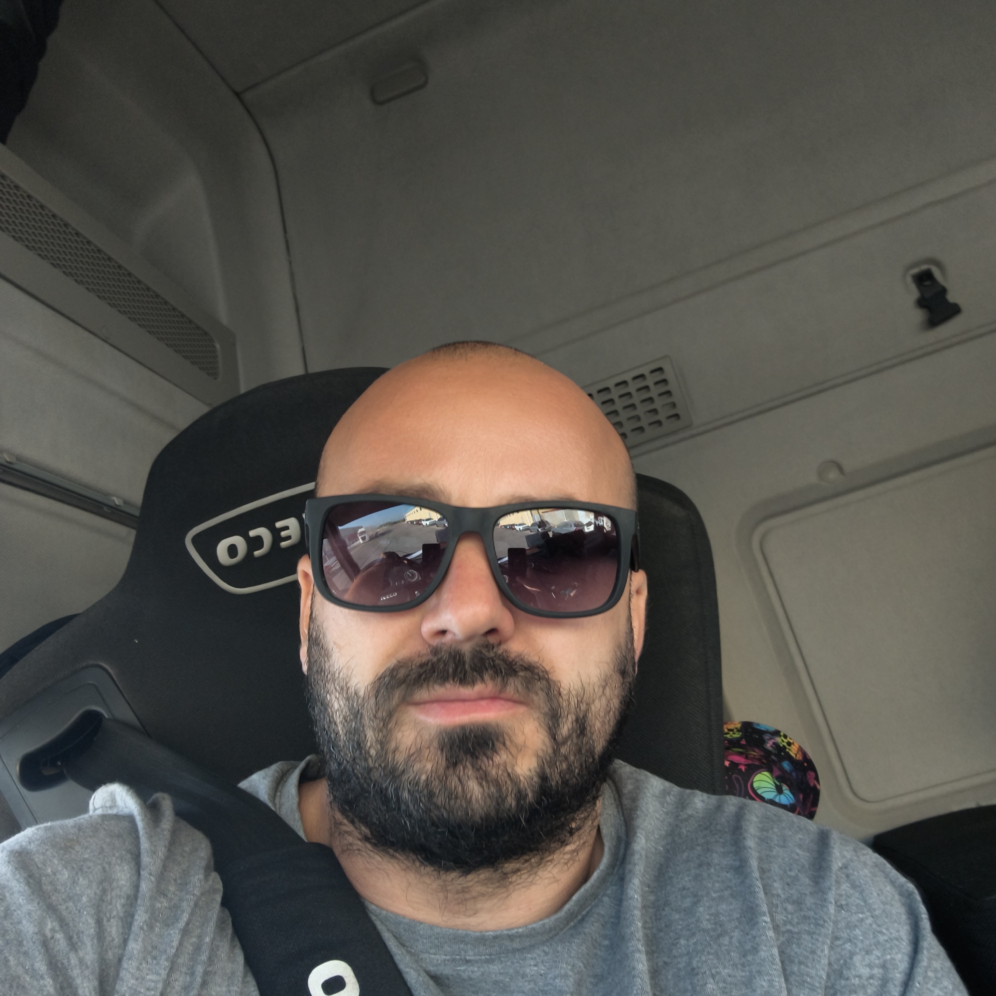 A bald man with a full beard, wearing a grey t-shirt and dark rectangular sunglasses, is captured in a close-up selfie. He is seated inside what appears to be the cab of a commercial vehicle, likely a truck, confirmed by the "IVECO" logo visible (reversed) on the black headrest of the seat behind him. The man's face is mostly obscured by his sunglasses and beard, with a neutral expression on his visible lower face.
The lenses of his sunglasses reflect an outdoor scene, showing a paved area with several parked vehicles, possibly other trucks or cars, under a clear, bright sky, indicating it is daytime. He is wearing a seatbelt, with a black strap crossing his chest, and a white letter "O" (or part of a logo) is visible on the seatbelt material near his shoulder. The interior of the vehicle is characterized by light grey ceiling panels and vents. The overall setting places the man in Manises, Spain.